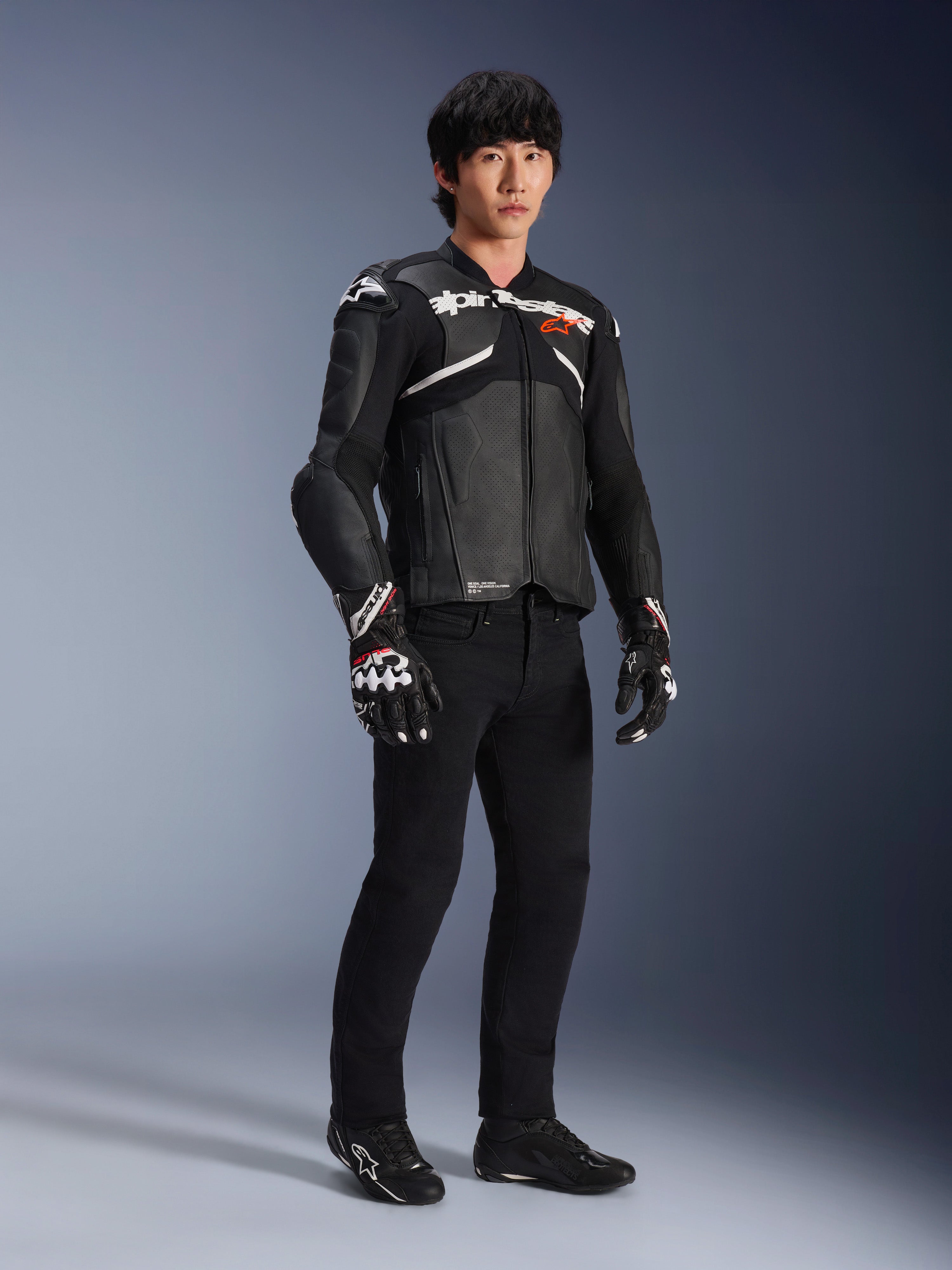 Alpinestars Atem V5 läderjacka, motorcykeljacka för racing, svart och vit, bärs med svarta byxor och matchande handskar, med vit Alpinestars-logotyp över bröstet och skyddande axelskydd.