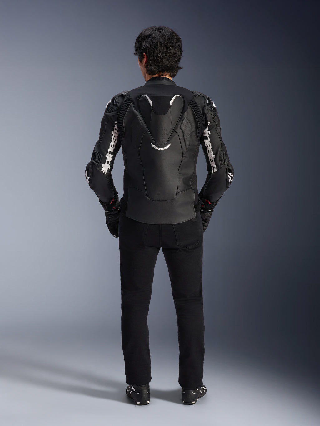 Alpinestars Atem V5 Läderjacka, motorcykeljacka för racing, svart och vit, vy bakifrån på förare, med aerodynamisk ryggpuckel och vit Alpinestars-logotyp på ärmar och rygg, teknisk läderkonstruktion, bärs med svarta byxor och MC-stövlar
