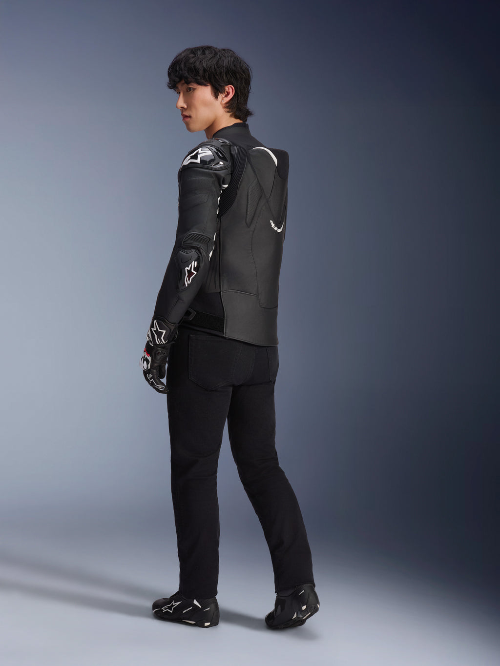 Manlig modell som bär Alpinestars Atem V5 läderjacka i svart och vitt, trekvarts vy bakifrån som visar den aerodynamiska ryggpuckeln och DFS RR-skydd, stylad med svarta byxor, racinghandskar och stövlar, professionell racingutrustning för motorcykel