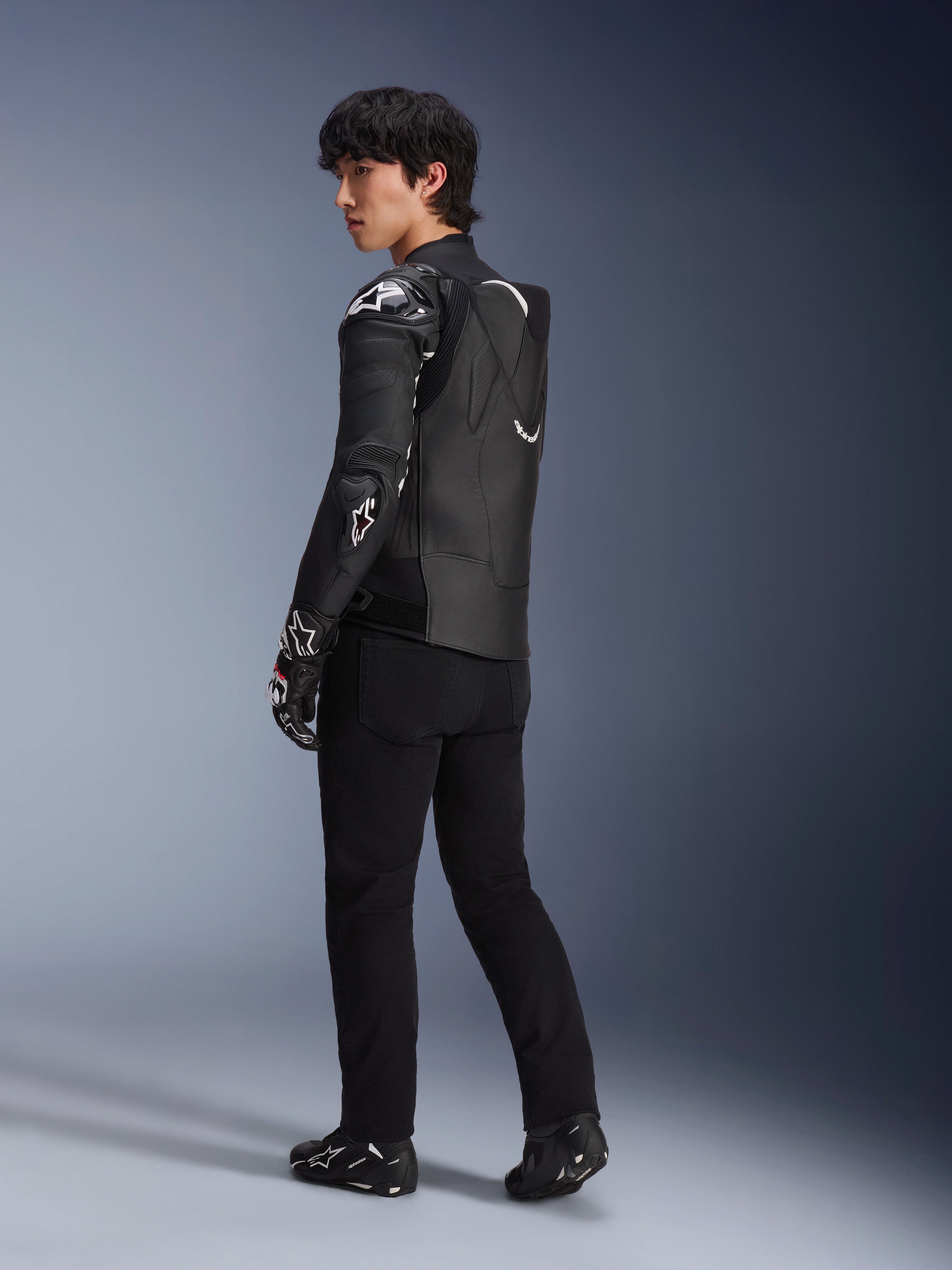 Manlig modell som bär Alpinestars Atem V5 läderjacka i svart och vitt, trekvarts vy bakifrån som visar den aerodynamiska ryggpuckeln och DFS RR-skydd, stylad med svarta byxor, racinghandskar och stövlar, professionell racingutrustning för motorcykel