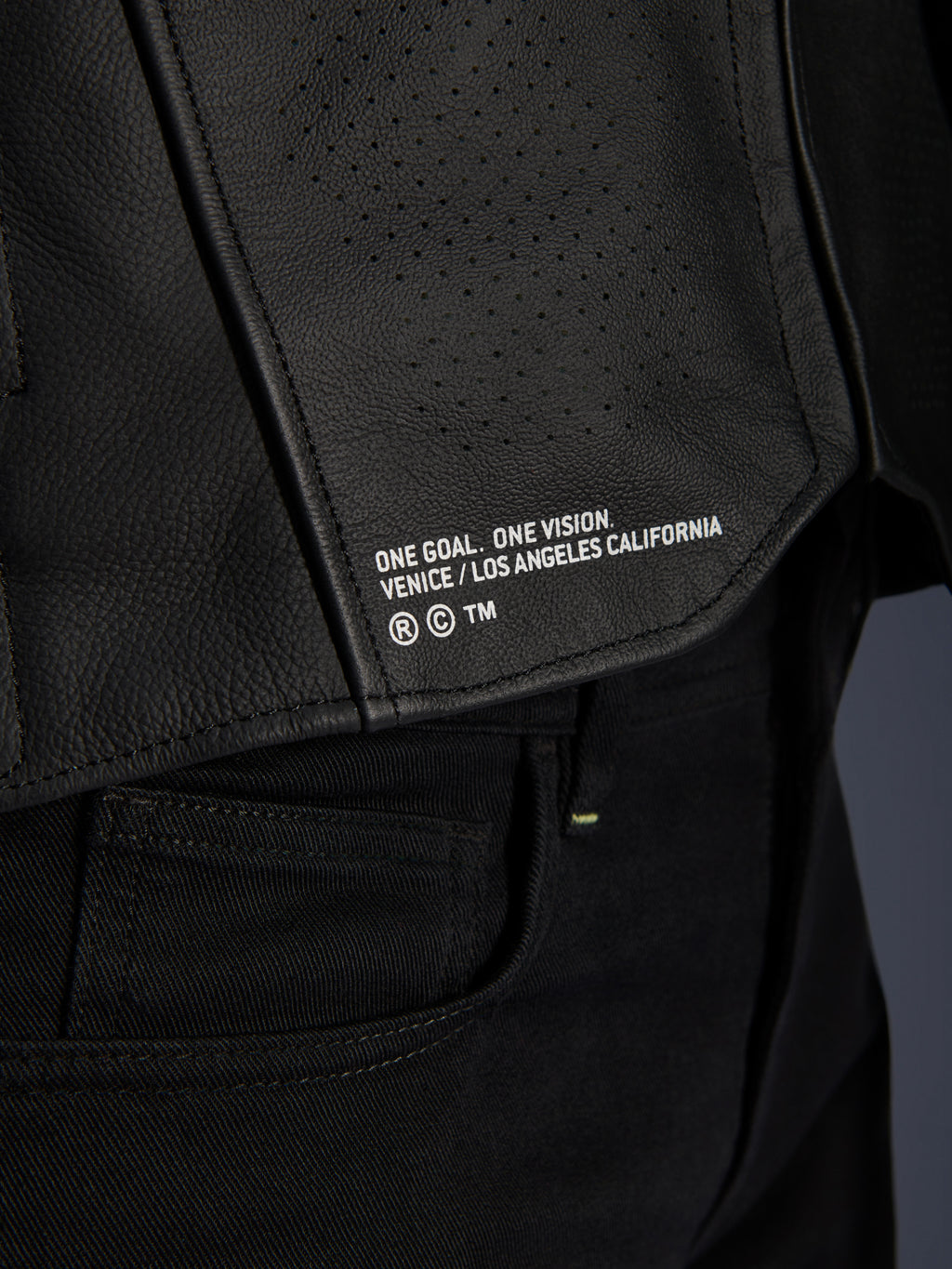 Närbild av Alpinestars Atem V5 läderjacka i svart, med den nedre ryggpanelen, 'ONE GOAL. ONE VISION. VENICE / LOS ANGELES CALIFORNIA'-tryck och perforerad läderstruktur.