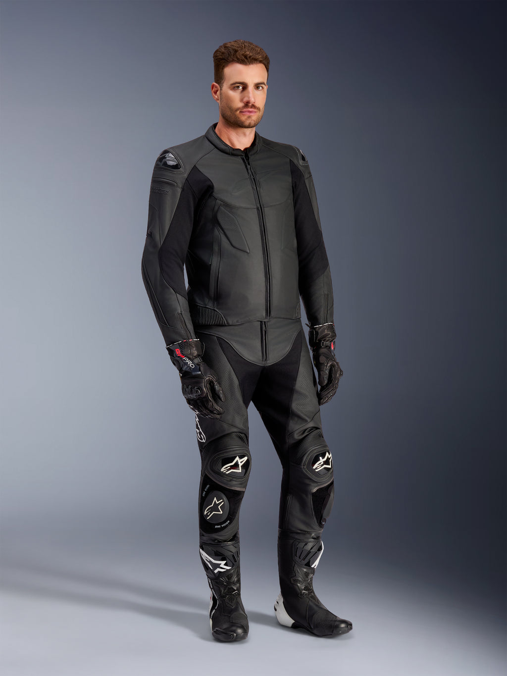 Male model wearing the Alpinestars Caliber Leather Jacket in Black, paired with matching black leather motorcycle pants, racing gloves, and boots, showcasing a complete sport road gear outfit.