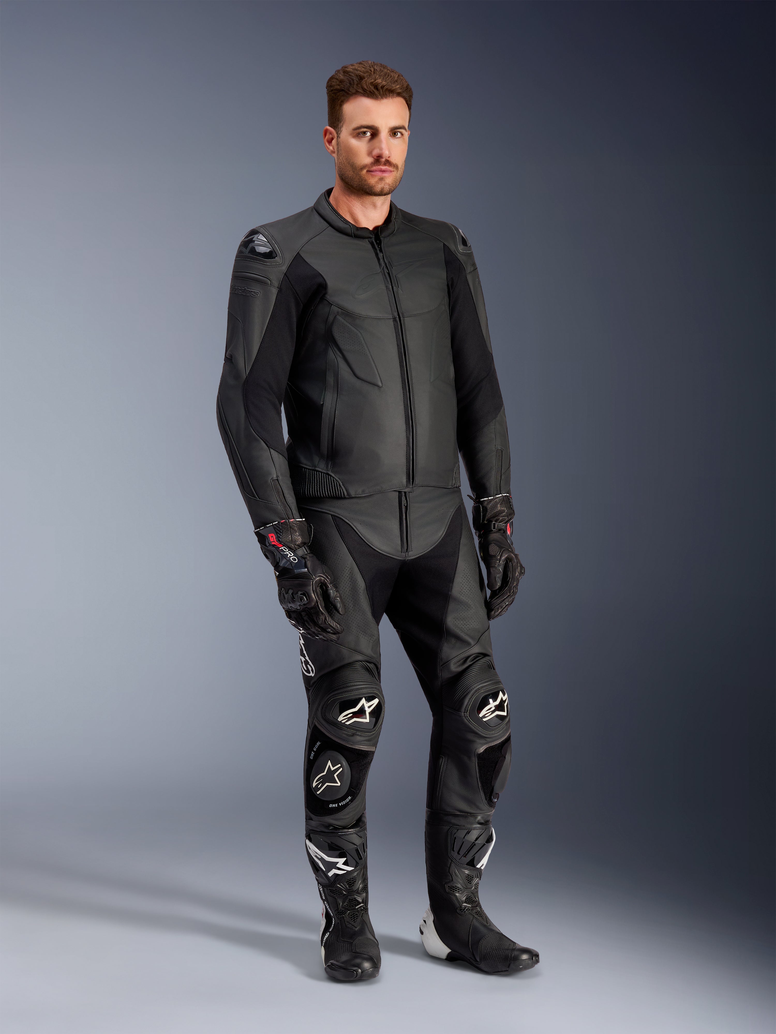 Male model wearing the Alpinestars Caliber Leather Jacket in Black, paired with matching black leather motorcycle pants, racing gloves, and boots, showcasing a complete sport road gear outfit.