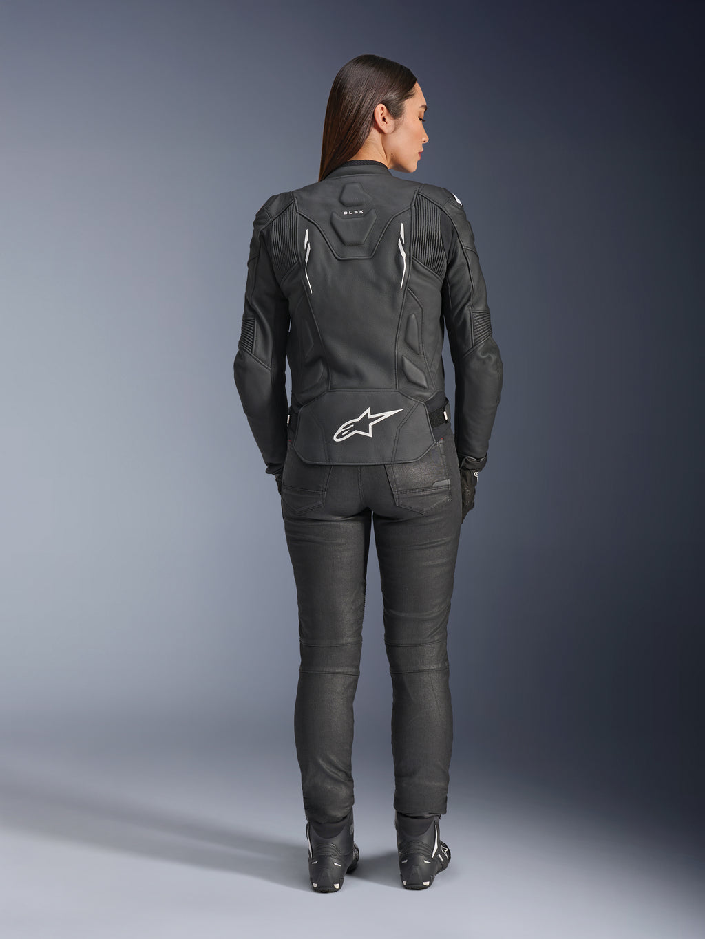 Alpinestars Stella Dusk läderjacka för dam, svart, sedd bakifrån med vit Alpinestars-logotyp på nedre delen av ryggen och 'DUSK'-märkning på övre delen av ryggen, buren med svarta MC-byxor och stövlar, framhäver den konturerade dampassformen och den skyddande läderkonstruktionen.