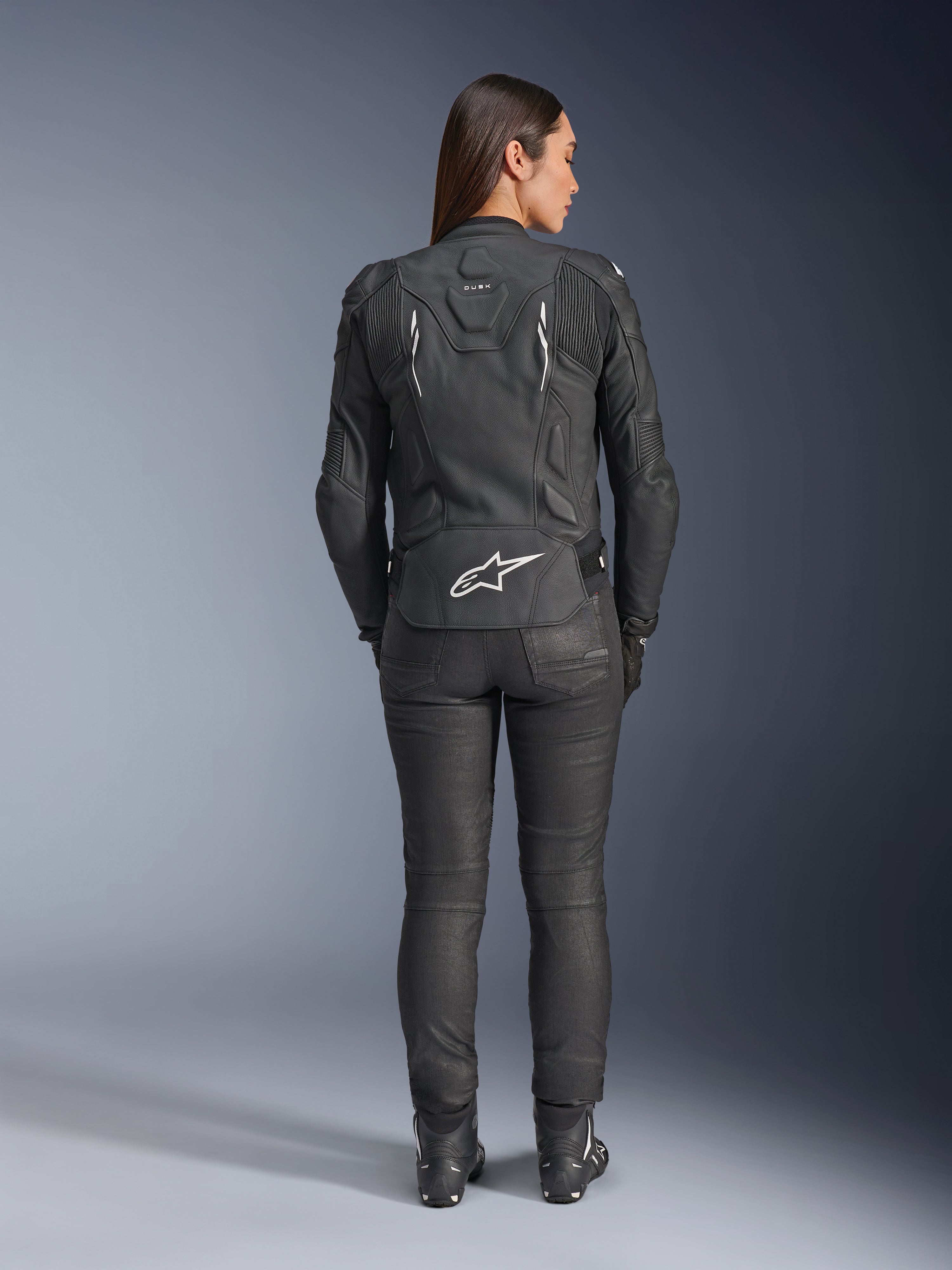 Alpinestars Stella Dusk läderjacka för dam, svart, sedd bakifrån med vit Alpinestars-logotyp på nedre delen av ryggen och 'DUSK'-märkning på övre delen av ryggen, buren med svarta MC-byxor och stövlar, framhäver den konturerade dampassformen och den skyddande läderkonstruktionen.