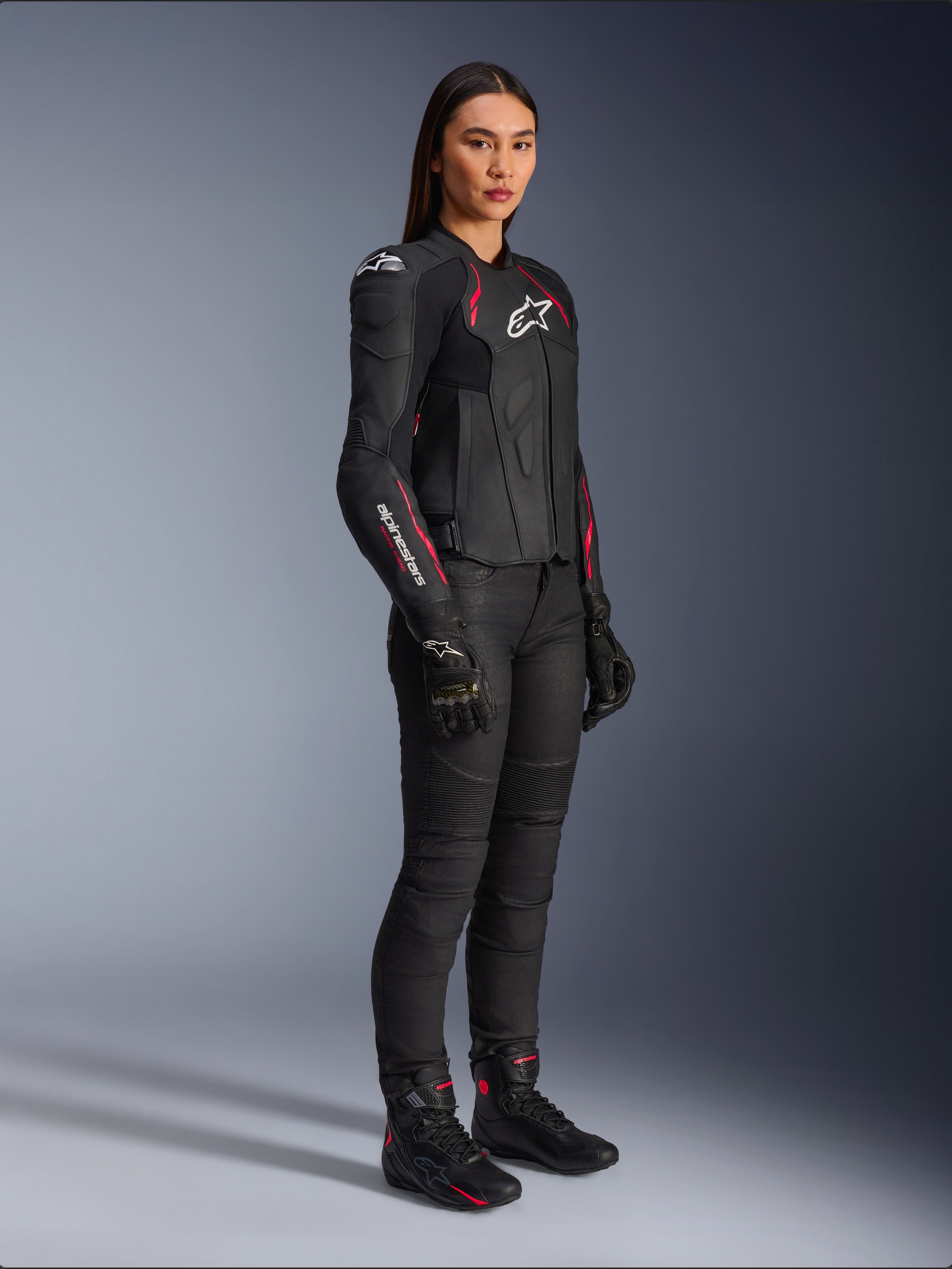 Alpinestars Stella Dusk skinnjacka för dam, svart, Diva Pink och vit, bärs av en kvinnlig modell med svarta motorcykelbyxor och stövlar, med en smidig läderkonstruktion, rosa detaljer och vita logotyper.