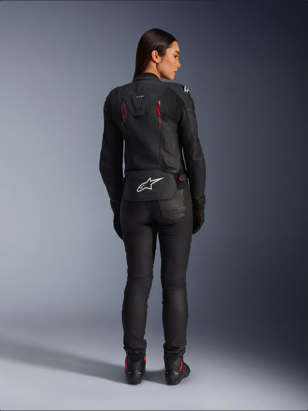 Alpinestars Stella Dusk läderjacka, vy bakifrån, svart läder med Diva Pink och vita detaljer, vit Alpinestars-logotyp på nedre delen av ryggen och rosa ventilationsdetaljer, bärs med svarta mc-byxor och stövlar, premium sportmotorcykelutrustning för damer