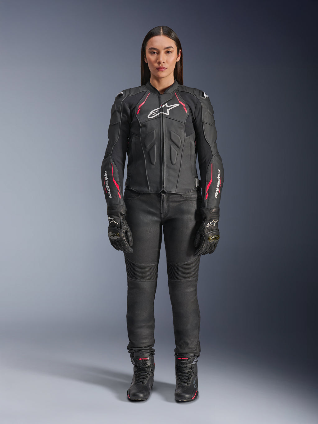 En kvinna som bär Alpinestars Stella Dusk läderjacka i Black Diva Pink White, med en vit logotyp på bröstet och rosa detaljer på axlarna och ärmarna, kombinerat med svarta motorcykelbyxor, handskar och stövlar.