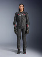 En kvinna som bär Alpinestars Stella Dusk läderjacka i Black Diva Pink White, med en vit logotyp på bröstet och rosa detaljer på axlarna och ärmarna, kombinerat med svarta motorcykelbyxor, handskar och stövlar.