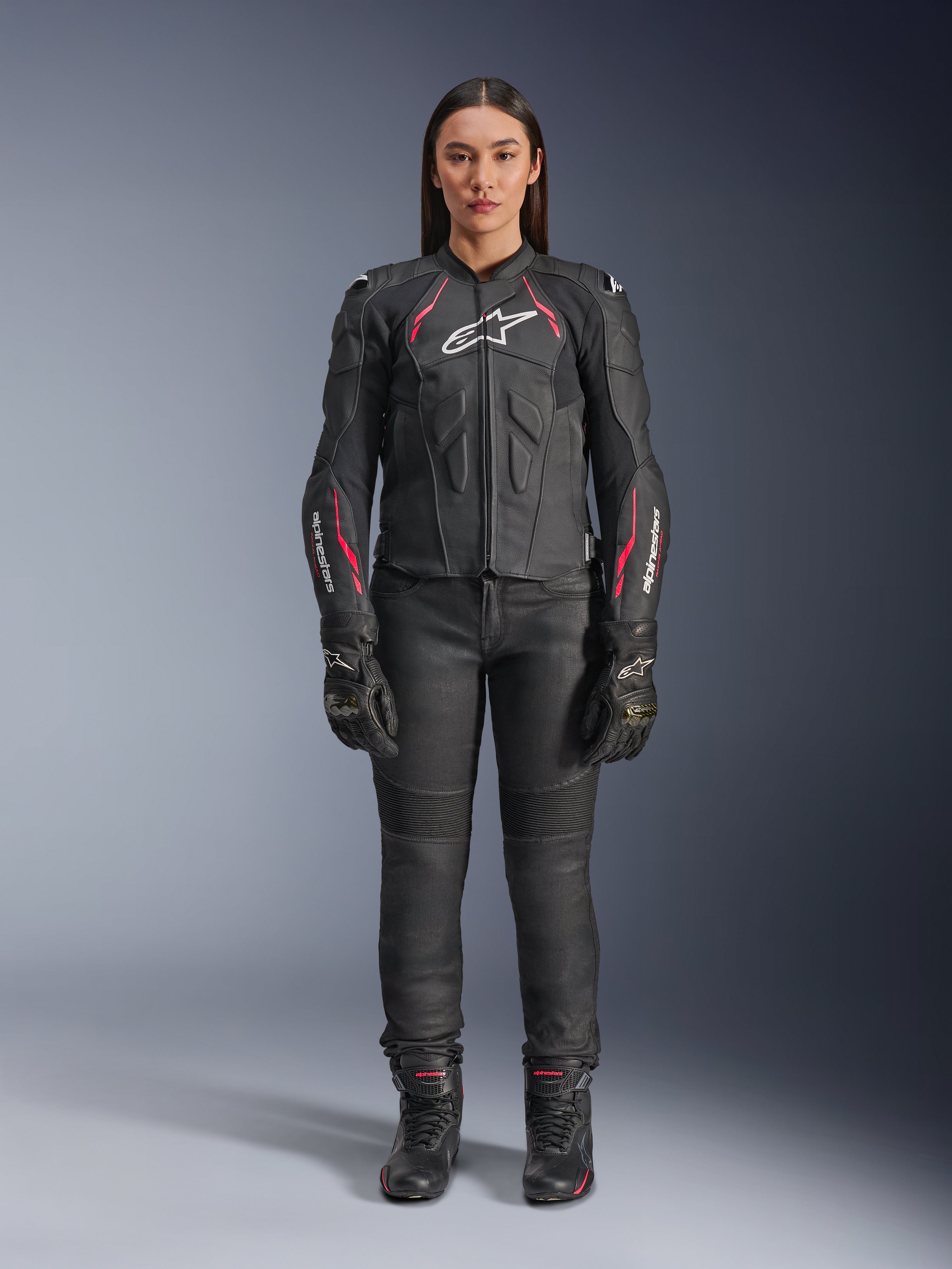 En kvinna som bär Alpinestars Stella Dusk läderjacka i Black Diva Pink White, med en vit logotyp på bröstet och rosa detaljer på axlarna och ärmarna, kombinerat med svarta motorcykelbyxor, handskar och stövlar.