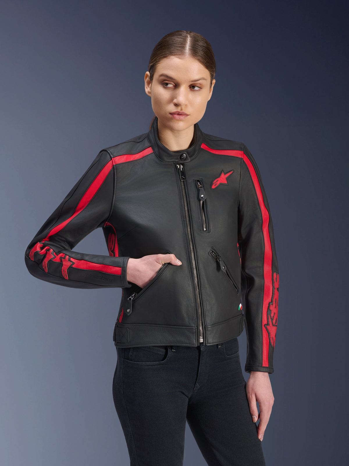 Alpinestars Stella Dyno läderjacka, motorcykeljacka för dam, svart och röd (Black Haute Red), bärs av en modell med svarta jeans, röda racingränder på axlar och ärmar med tydlig logotyp, premiumläderkonstruktion, heritage-inspirerad design för urban körning