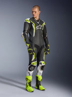 Alpinestars GP Tech V5 One Piece Leather Suit, svart, vit och neongul, bärs av en modell med matchande racinghandskar och stövlar, professionell motorcykelutrustning med aerodynamisk prestanda och inbyggda skydd.