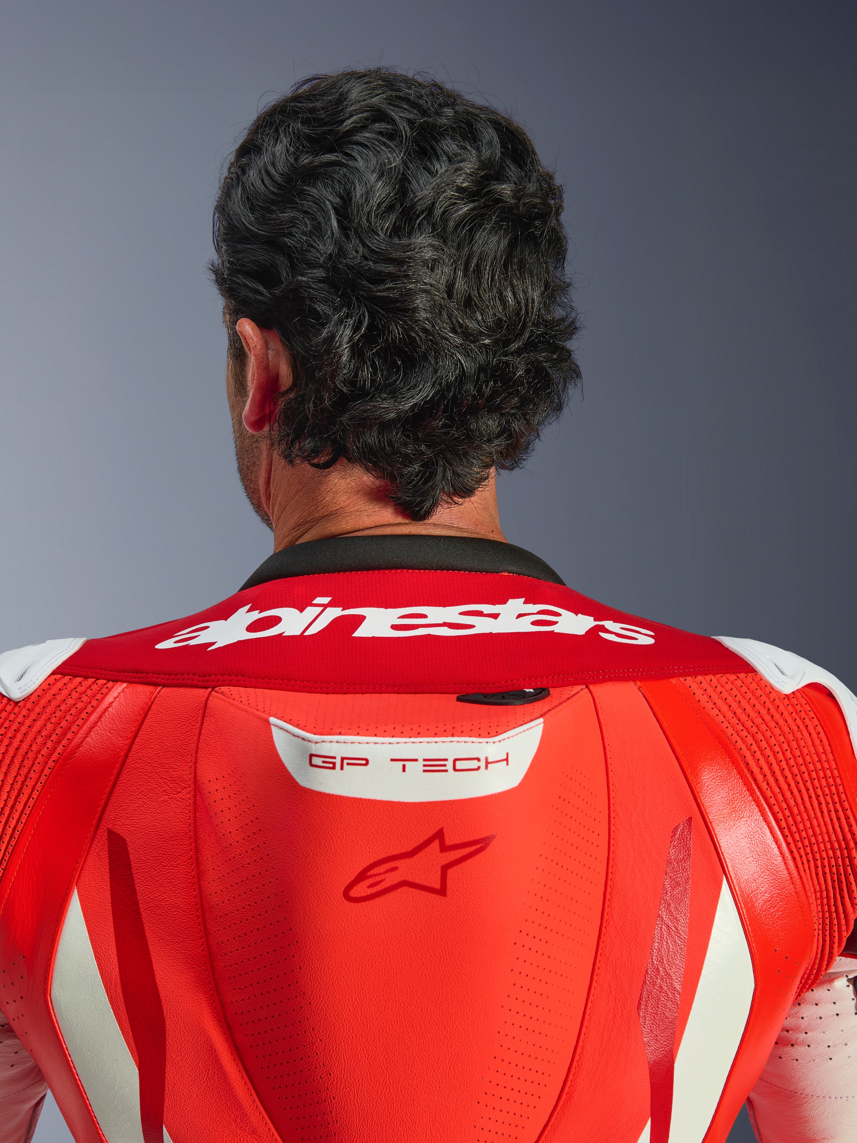 Close-up of the upper back and neck of the GP Tech V5 One Piece Leather Suit, Red Fluo and Bright Red, featuring white Alpinestars wordmark on the collar and GP TECH logo on the aerodynamic hump, worn by a rider.