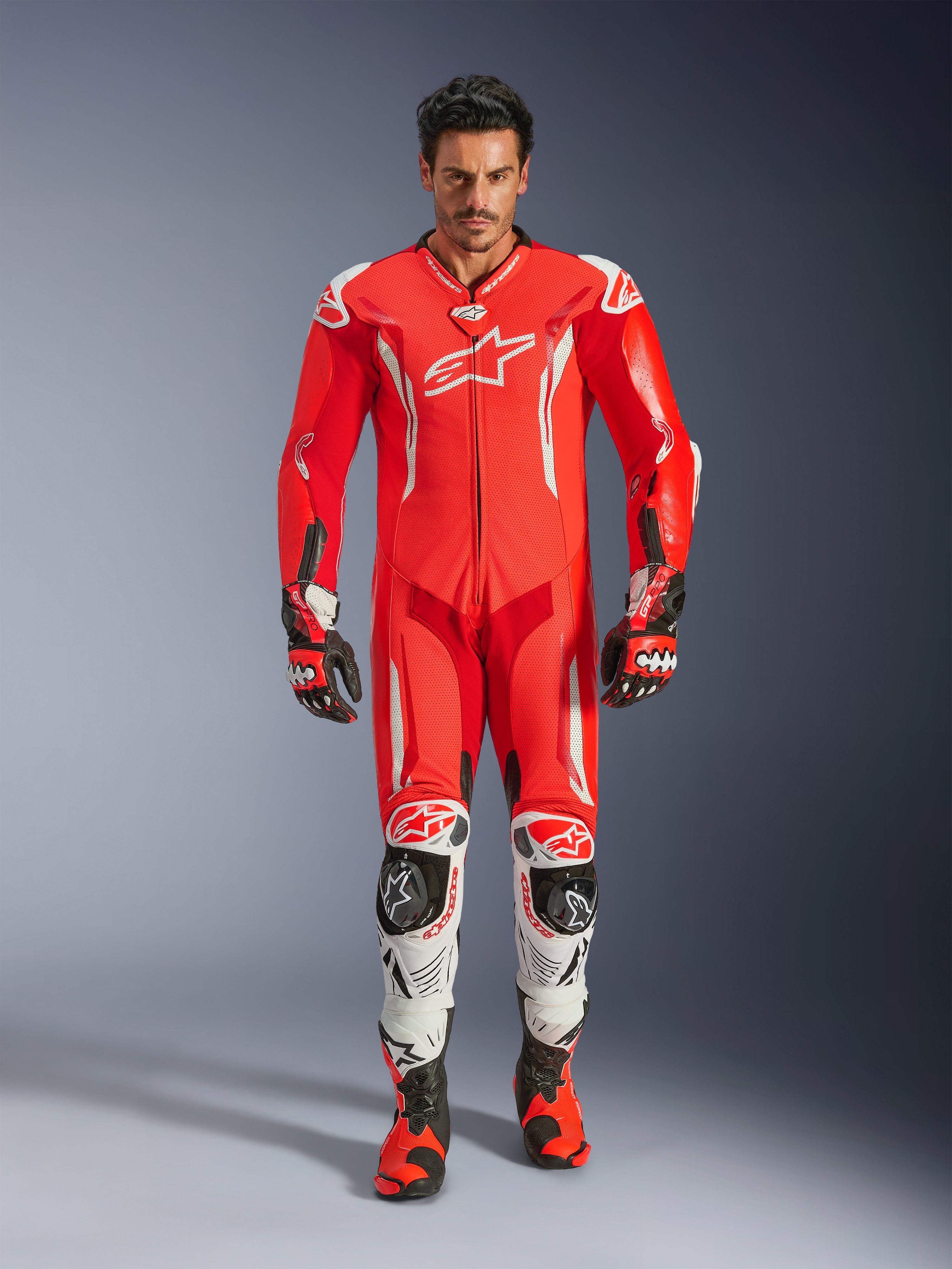 Alpinestars GP Tech V5 racingoverall i ett stycke, professionell racingoverall i Red Fluo och Bright Red med vita accenter, bärs av en förare med matchande stövlar och handskar, utrustad med en aerodynamisk puckel, premium läderkonstruktion och avancerat skydd för bankörning