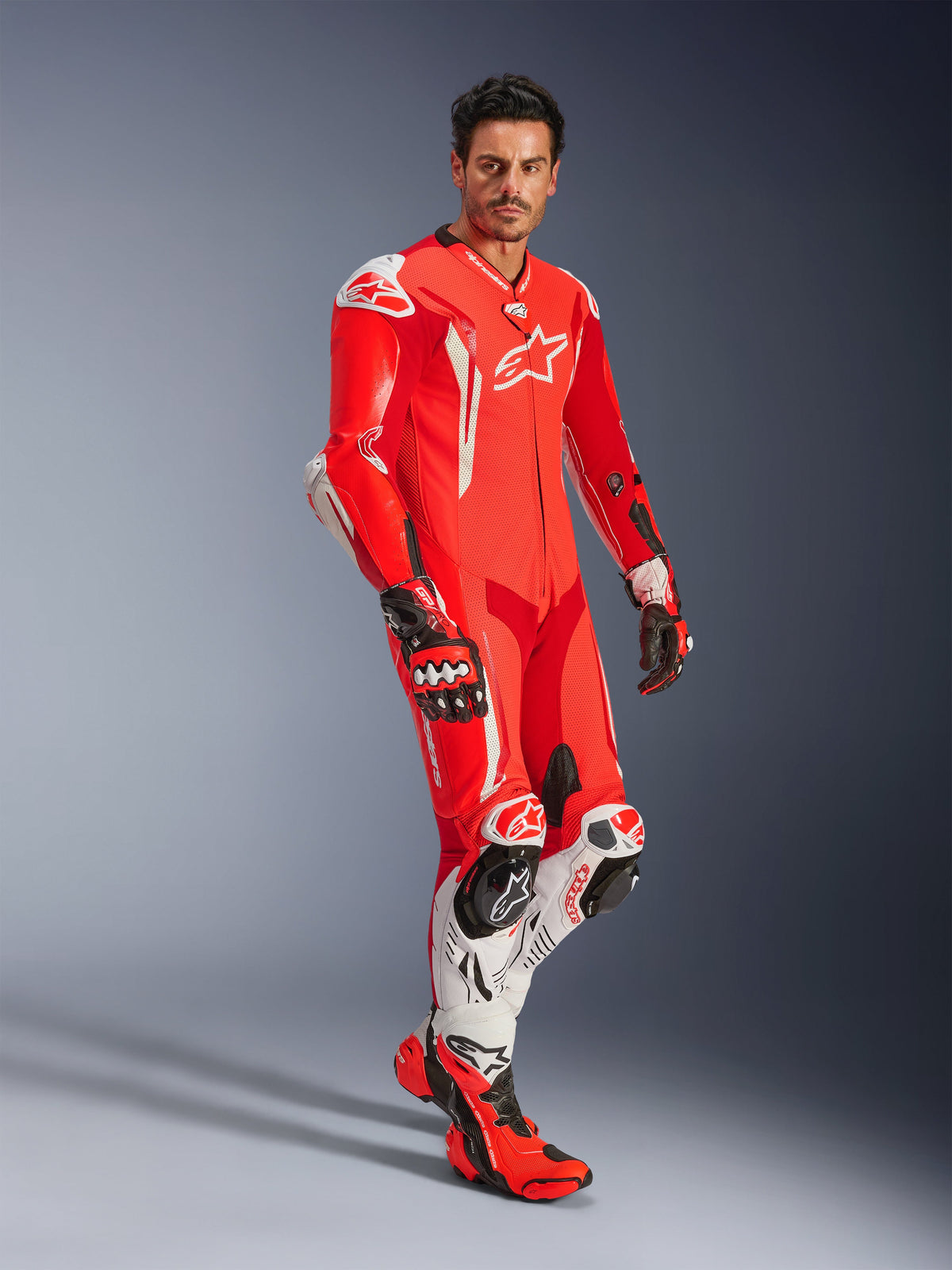 Man som bär Alpinestars GP Tech V5 endelat skinnställ i Red Fluo, Bright Red och vitt, koordinerat med racinghandskar och stövlar, professionell racingutrustning för motorcykel med aerodynamisk puckel och integrerat skydd.