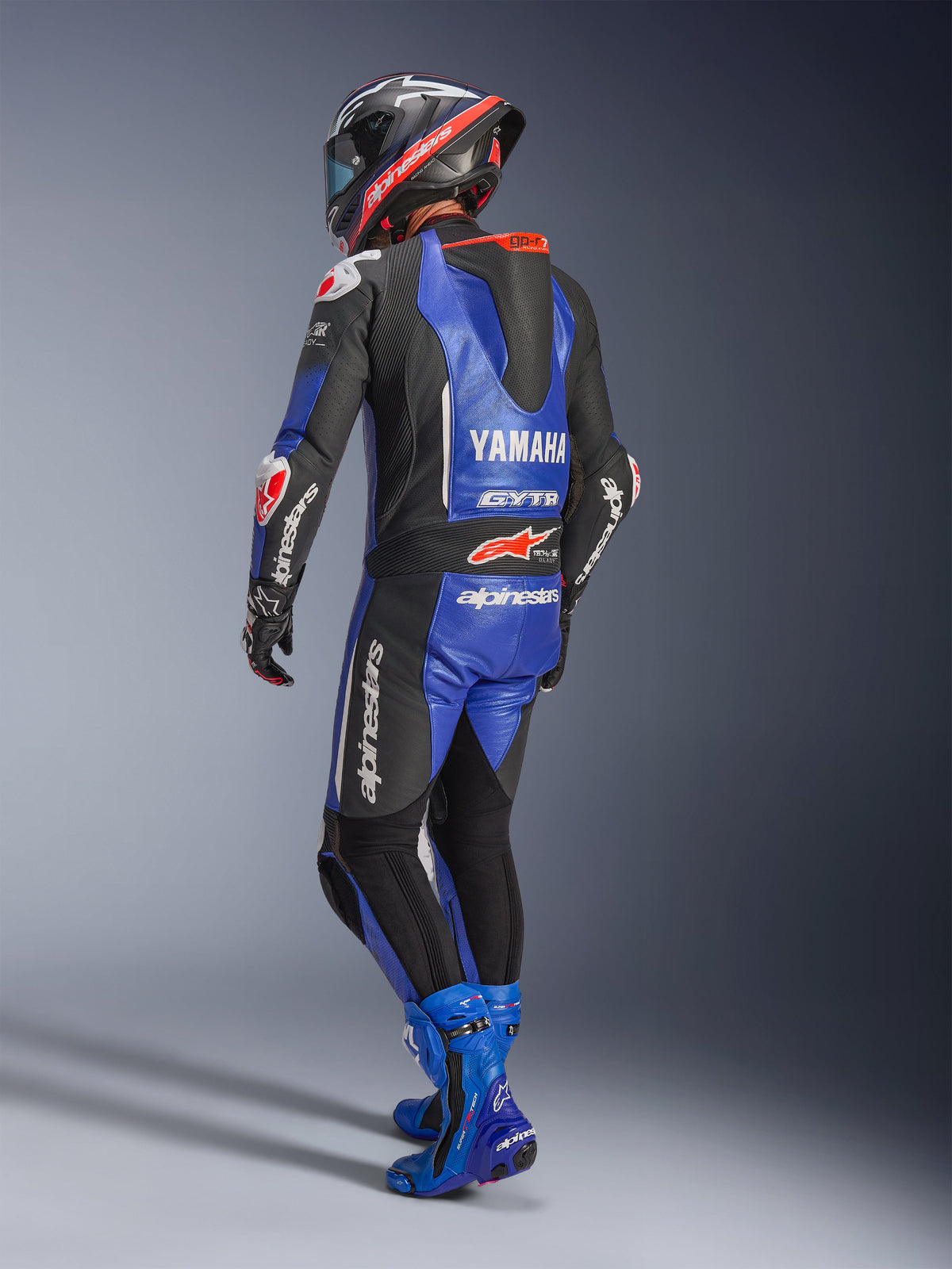Alpinestars Yamaha GP-R7 endels läderdräkt, metallisk blå, svart och vit, vy bakifrån som visar aerodynamisk puckel och Yamaha-logotyp, bärs med matchande blå racingstövlar och hjälm, professionellt skinnställ för banracing