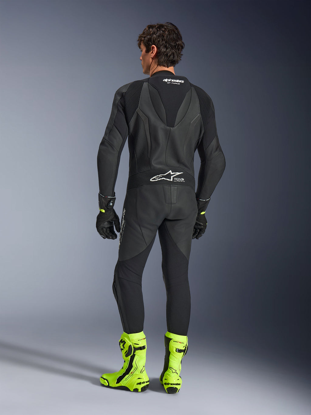 Alpinestars GP Force V2 Leather Suit - 1Piece, Black, back view worn by a model, featuring an aerodynamic speed hump and Tech-Air Ready branding, styled with black gloves and bright fluorescent yellow racing boots, professional track equipment
