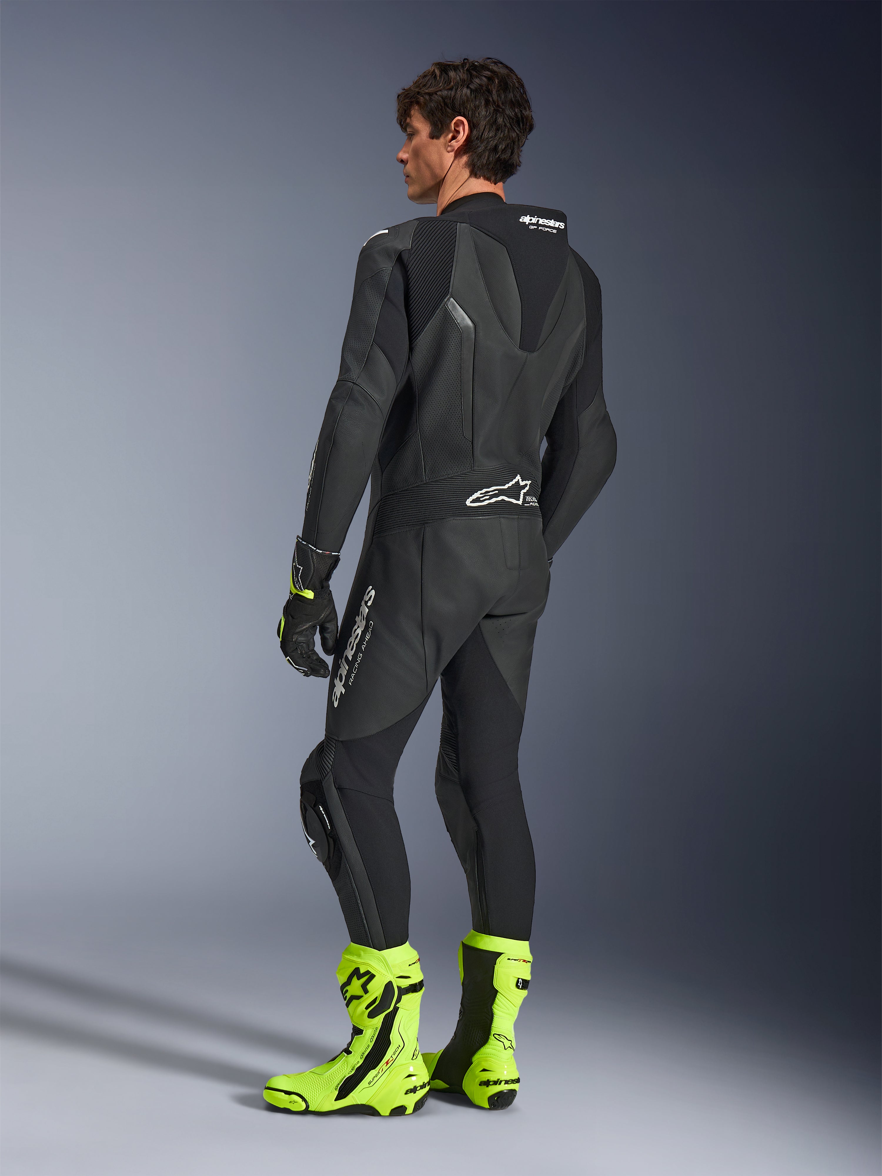 Alpinestars GP Force V2 Leather Suit, 1-Piece Black motorcycle racing suit, rear view showing aerodynamic speed hump and perforated leather, worn with neon yellow racing boots and black gloves, professional track-ready equipment