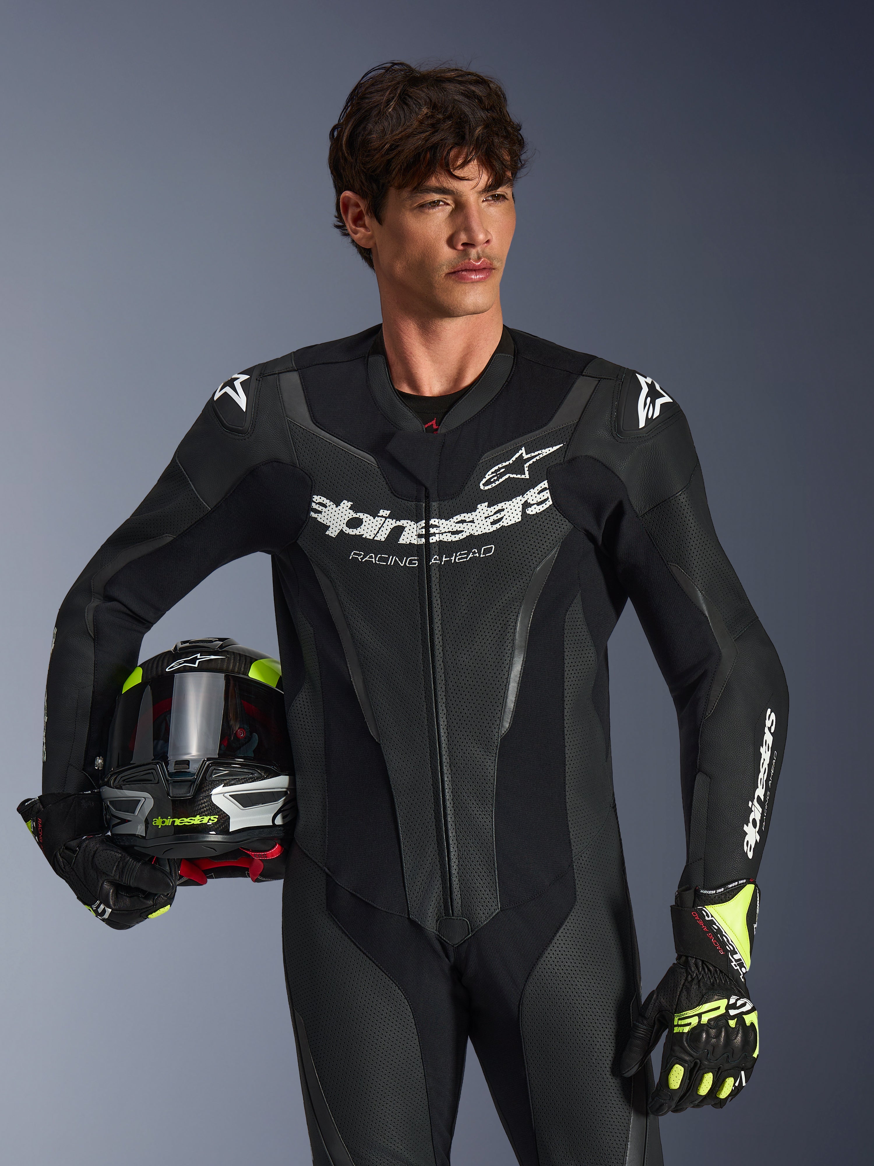 Alpinestars GP Force V2 Leather Suit, 1-Piece black motorcycle racing suit with perforated leather panels and white logo branding, worn by a model holding a helmet and matching gloves, studio lighting