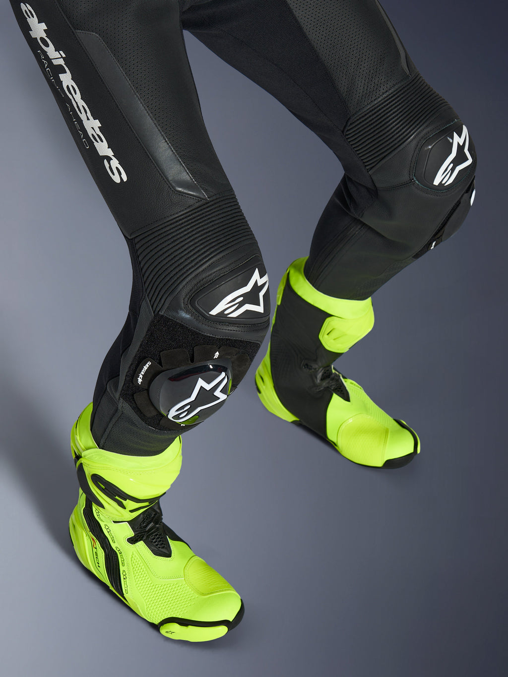 Alpinestars GP Force V2 Leather Suit, 1-Piece Black Leather Suit, close-up of legs and knees featuring white logo sliders, accordion stretch panels, worn with bright neon yellow racing boots, high-performance motorcycle racing gear