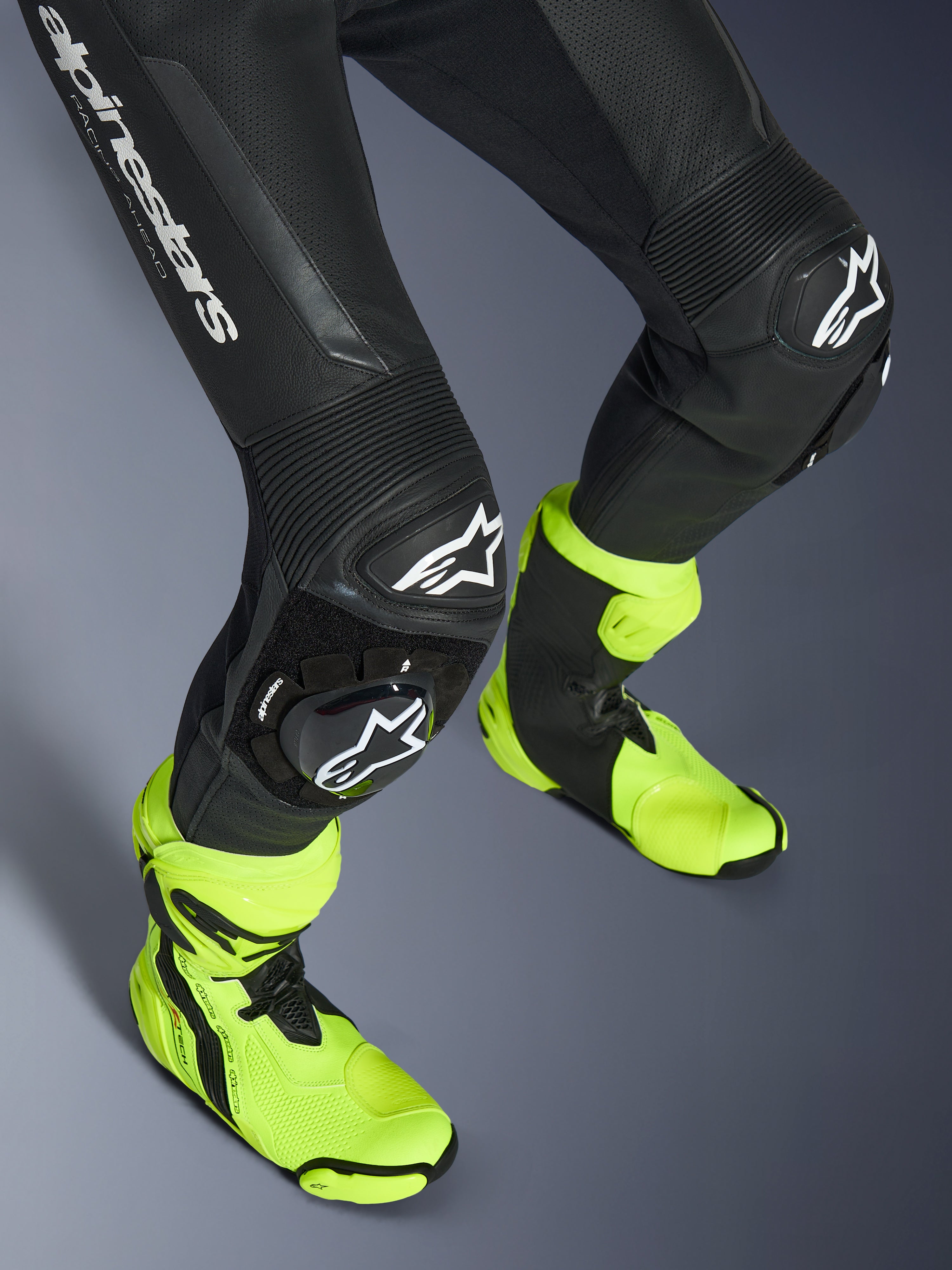 Alpinestars GP Force V2 Leather Suit, 1-Piece Black Leather Suit, close-up of legs and knees featuring white logo sliders, accordion stretch panels, worn with bright neon yellow racing boots, high-performance motorcycle racing gear