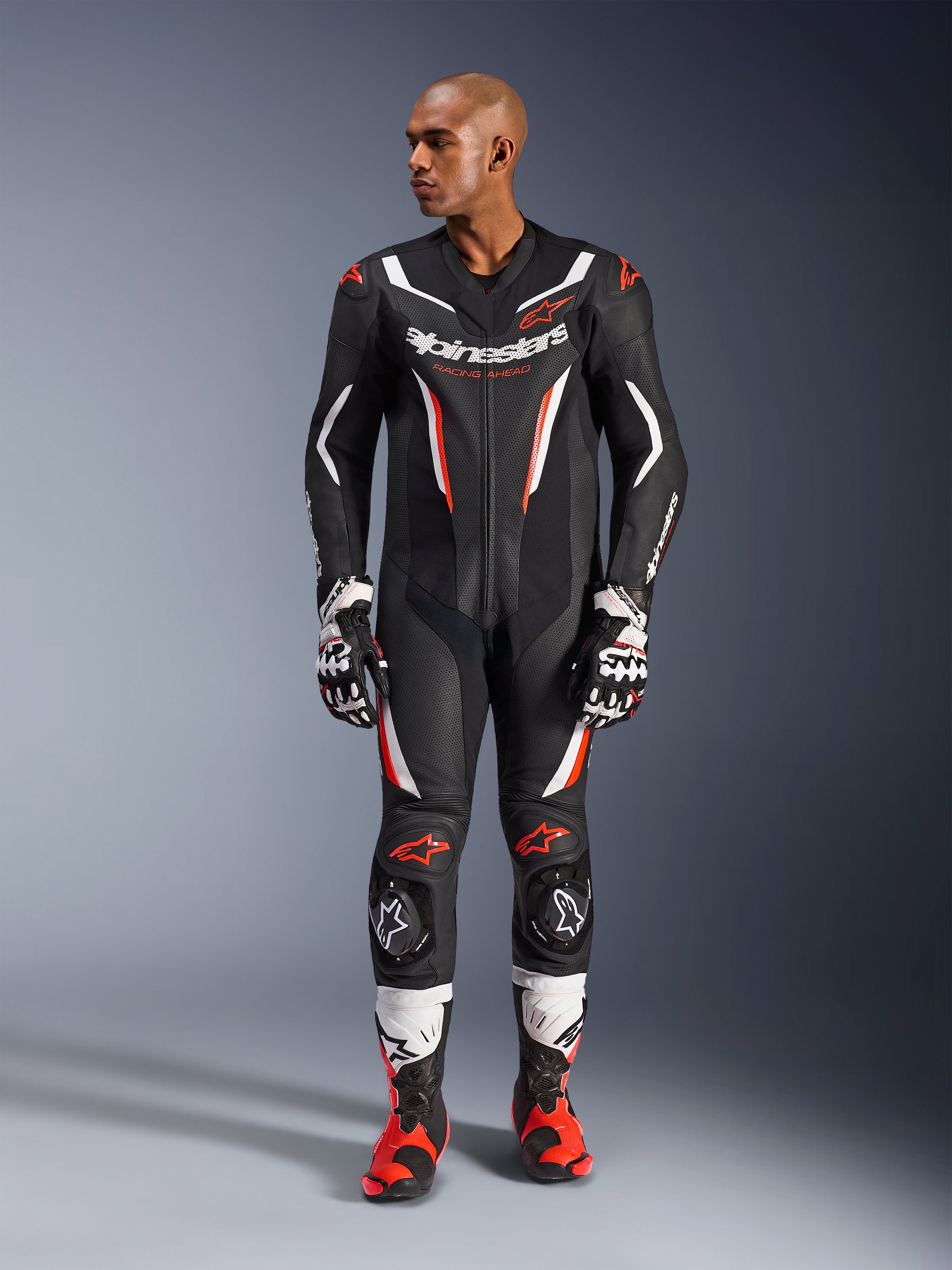 Alpinestars GP Force V2 skinnställ - 1 del, svart, vit och röd fluo, helkroppsbild av manlig modell i professionell racingutrustning för motorcykel med matchande handskar och stövlar, med perforerade läderpaneler och skyddande knäpuckar.