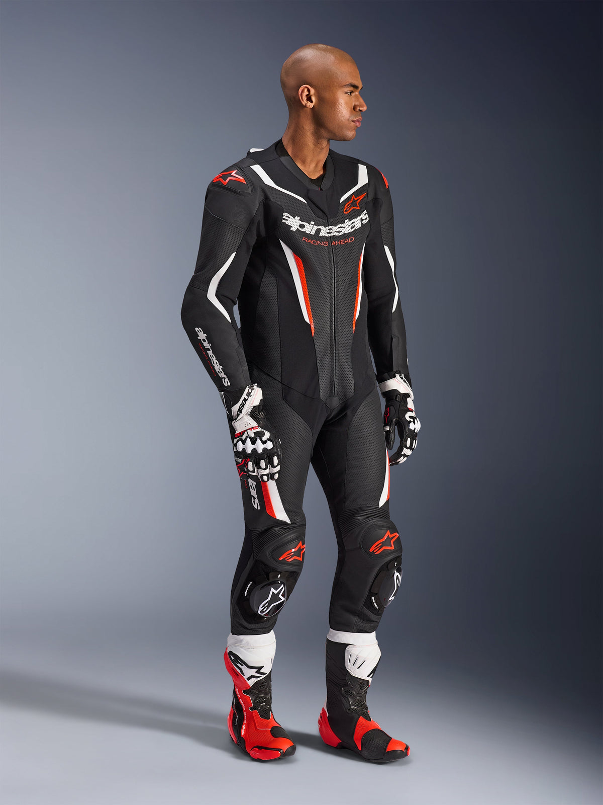 Modell som bär Alpinestars GP Force V2 skinnställ i ett stycke i Black White Red Fluo, högpresterande racingutrustning med perforerade läderpaneler och DFS Lite-skydd, stylad med matchande röda och vita racingstövlar och handskar, professionell motorsportestetik.