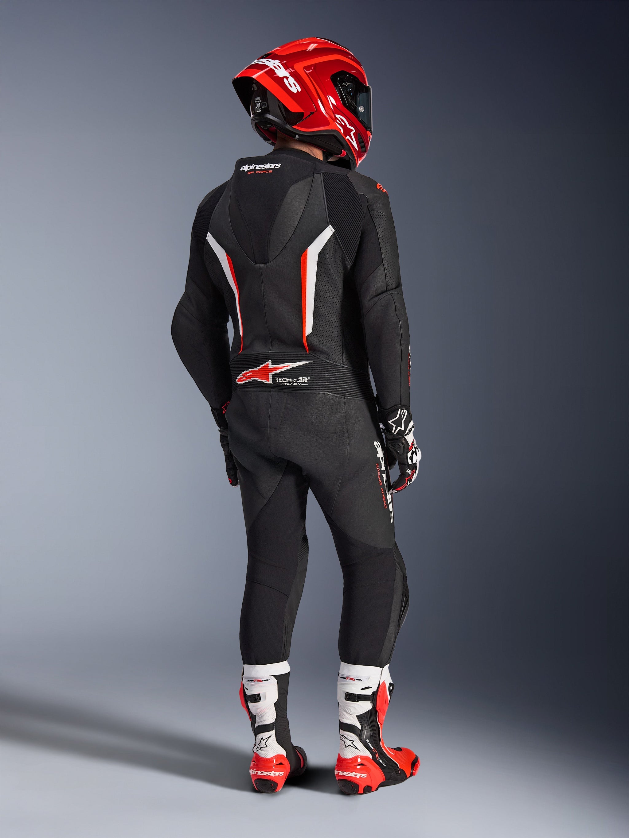 Rider seen from behind wearing the Alpinestars GP Force V2 1-Piece Leather Suit in Black, White, and Red Fluo, featuring an aerodynamic hump and perforated leather panels, paired with a red Alpinestars helmet, racing gloves, and red and white motorcycle boots, professional track racing gear.