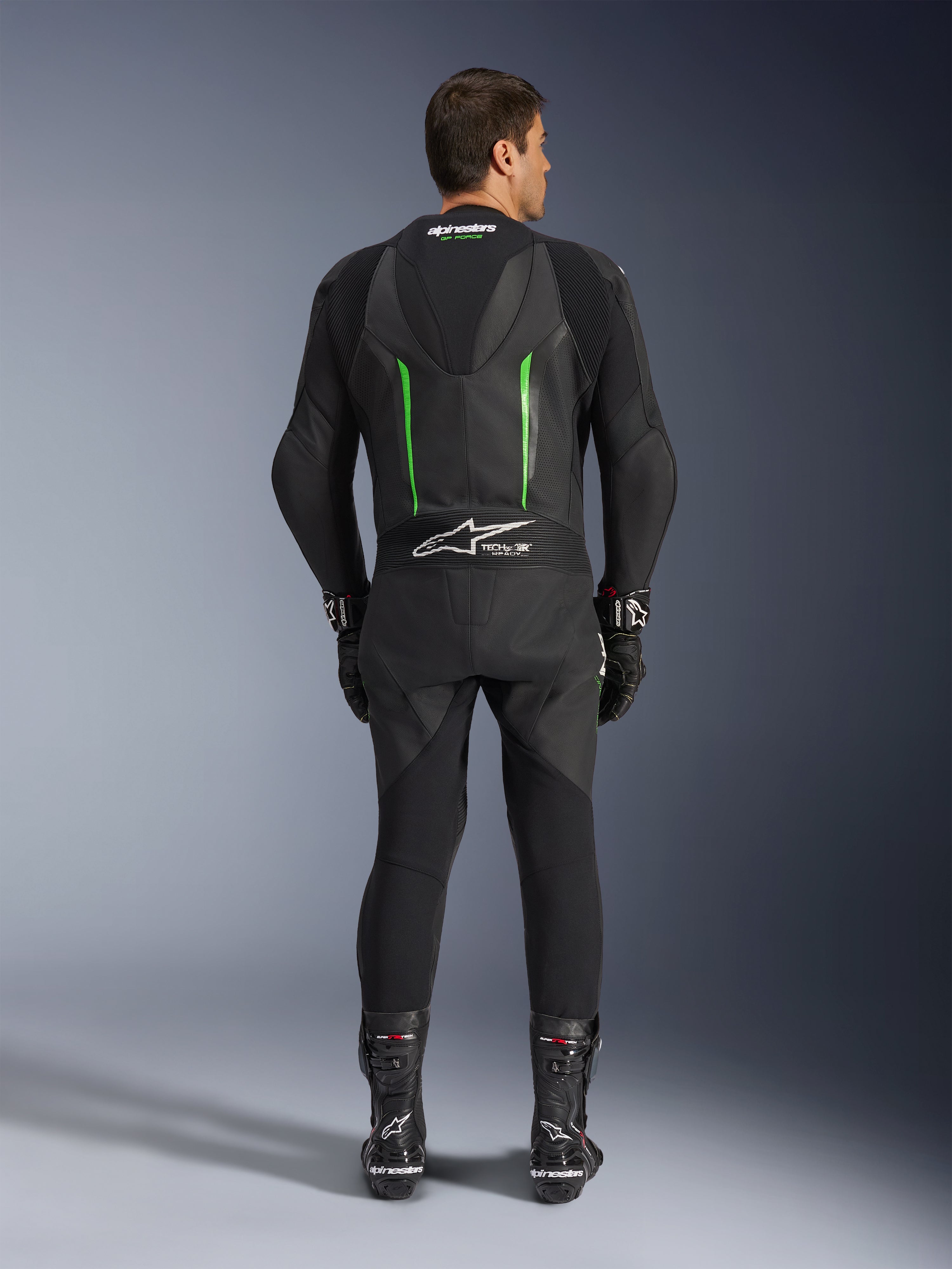 Vy bakifrån av Alpinestars GP Force V2 1-Piece skinnställ i svart och grönt, med en aerodynamisk speedpuckel, perforerade paneler och Tech-Air Ready-märkning på nedre delen av ryggen, bärs med svarta racinghandskar och stövlar.