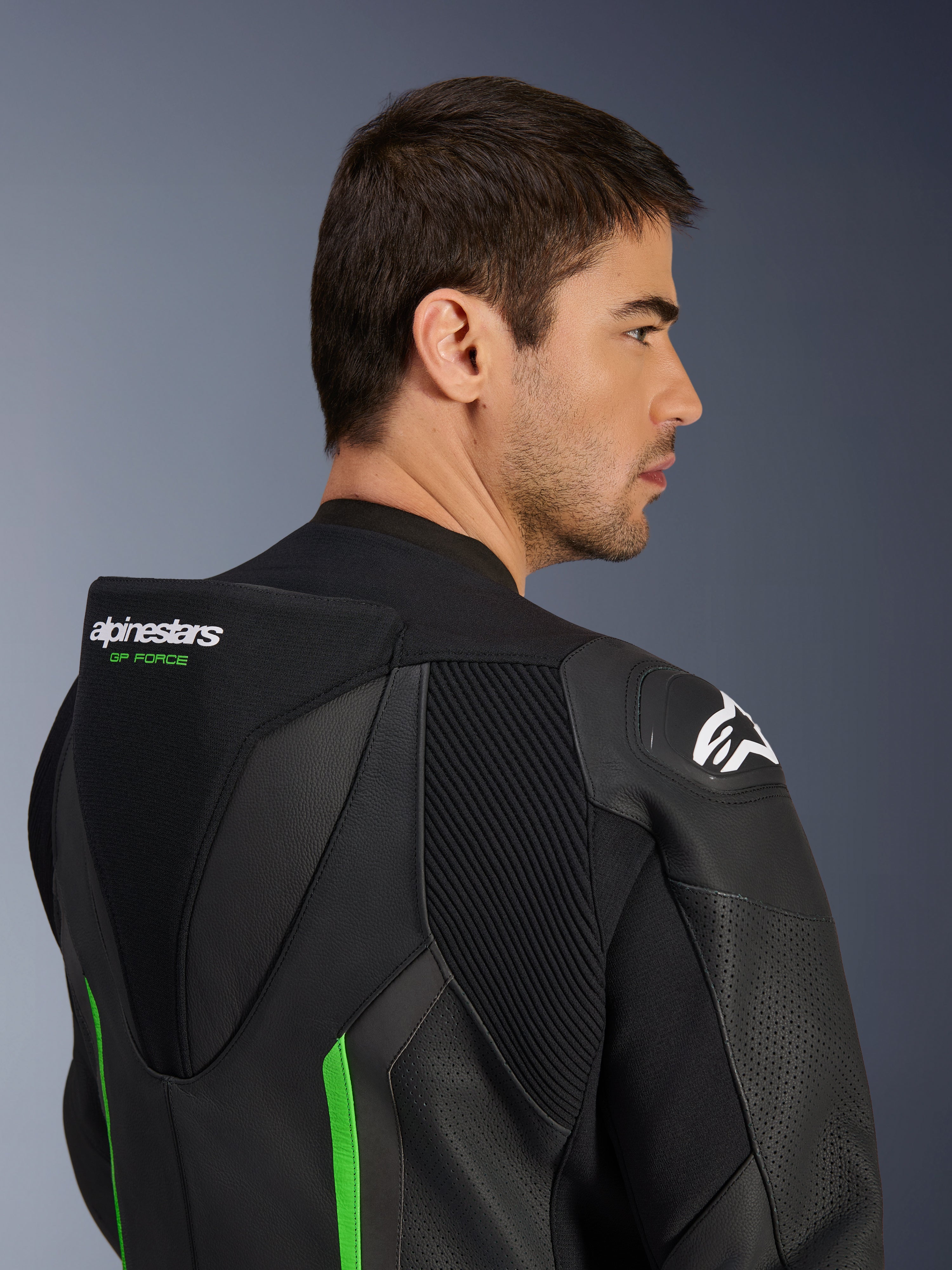 Närbild på övre ryggen och axeln på en manlig modell som bär Alpinestars GP Force V2 Leather Suit i svart och grönt, med den aerodynamiska puckeln med grön 'GP FORCE'-branding, en vit Astars-logotyp på axeln och dragspelspaneler.