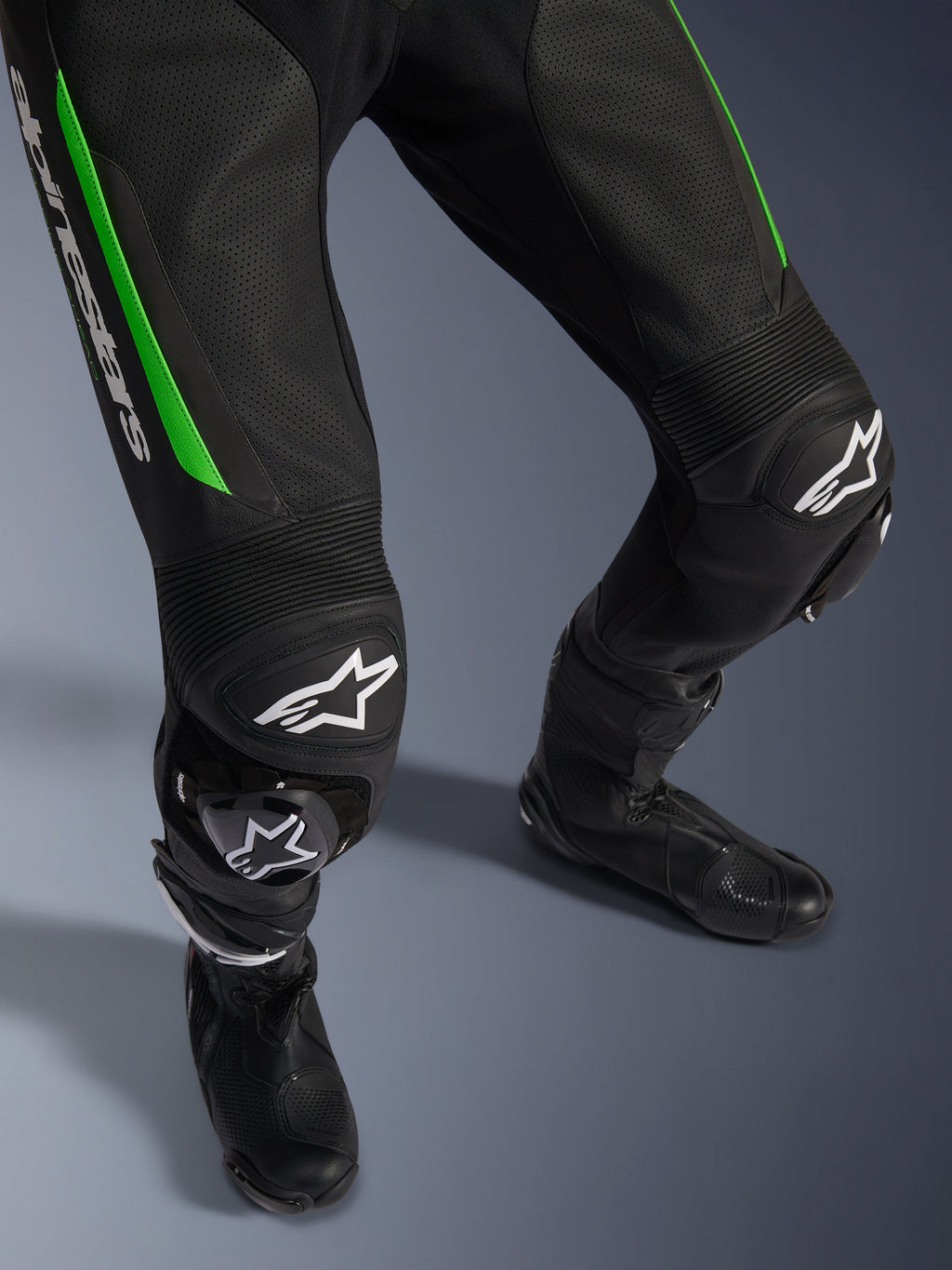 Alpinestars GP Force V2 läderställ - 1 del, närbild på ben i svart och grönt, med perforerad läderkonstruktion, dragspelspaneler i stretch vid knäna, vit Alpinestars-logotyp och integrerade knäpuckar, bärs med svarta racingstövlar