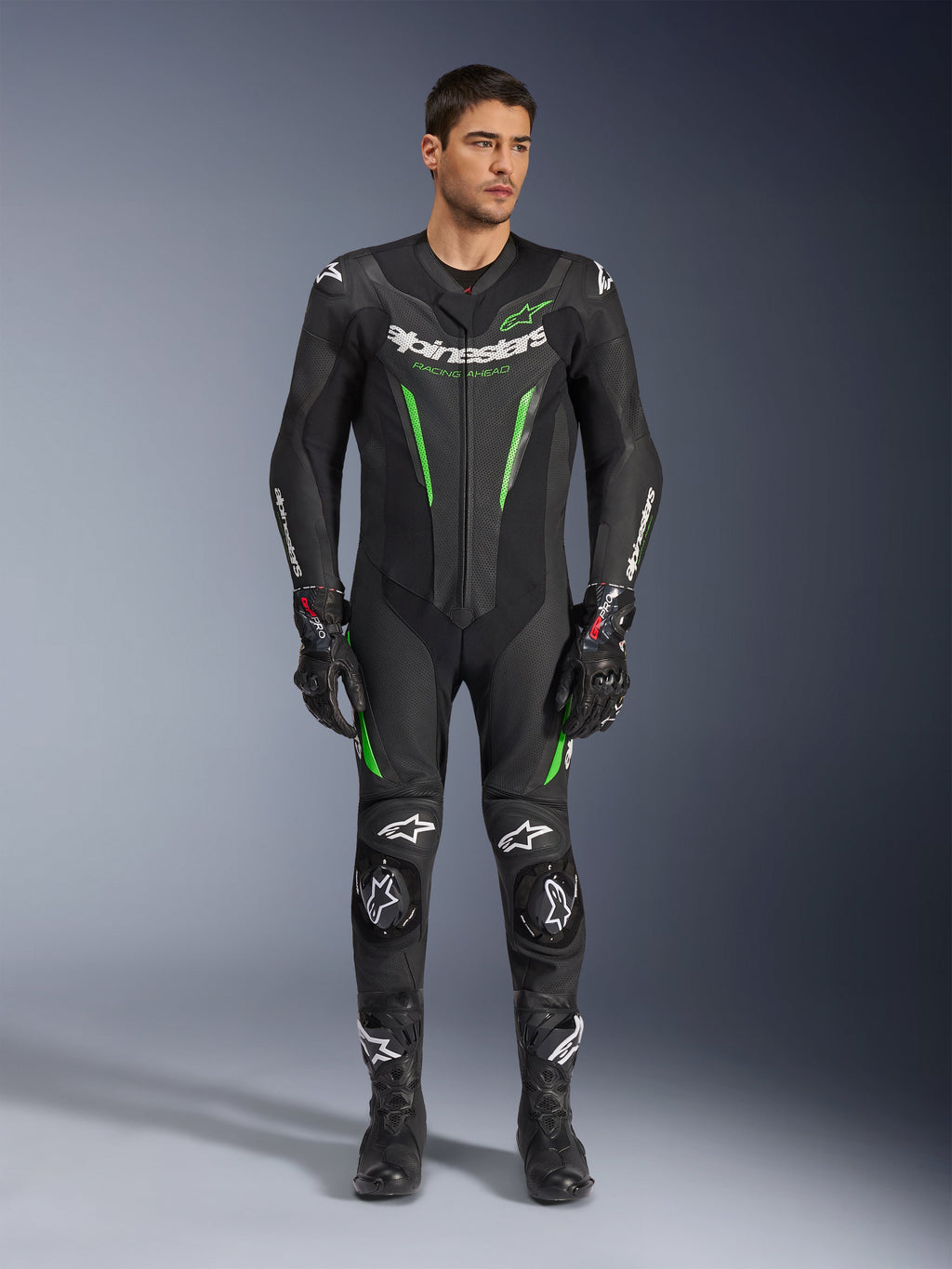 Alpinestars GP Force V2 Leather Suit - 1Piece, heldelat skinnställ för motorcykelracing, svart och grön, frontvy av en modell i stället med vit logotyp på bröstet och neongröna detaljer, med perforerat läder och externa axelskydd, kombinerat med svarta racinghandskar och stövlar