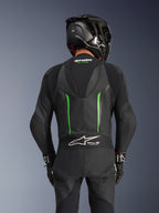 Baksida av en förare klädd i Alpinestars GP Force V2 Leather Suit i Black Green, med en aerodynamisk speedpuckel, Tech-Air Ready-märkning på dragspelspanelen i ländryggen och perforerad läderkonstruktion, kombinerat med en svart racinghjälm i kolfiber.