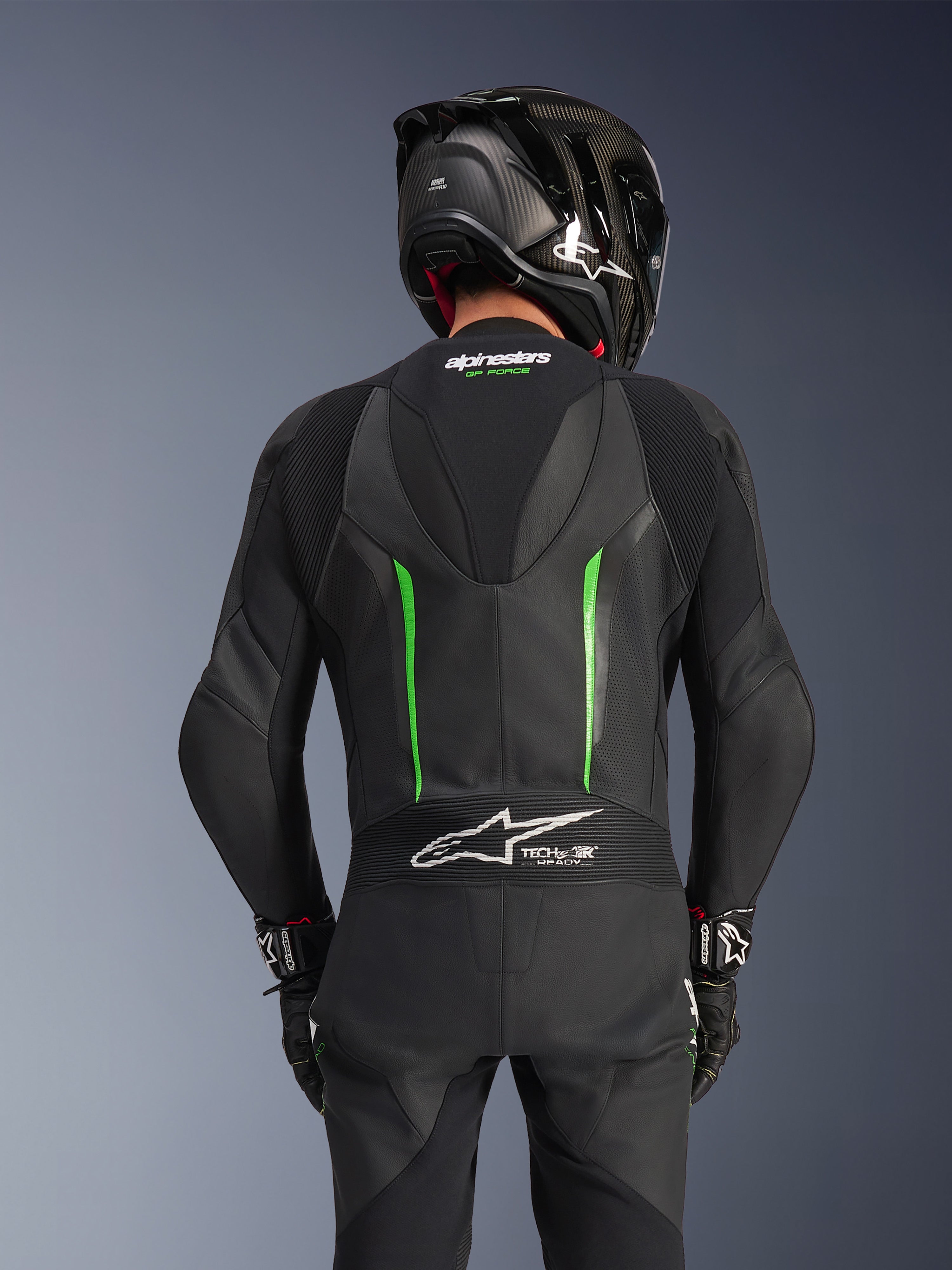 Baksida av en förare klädd i Alpinestars GP Force V2 Leather Suit i Black Green, med en aerodynamisk speedpuckel, Tech-Air Ready-märkning på dragspelspanelen i ländryggen och perforerad läderkonstruktion, kombinerat med en svart racinghjälm i kolfiber.