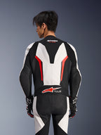 Alpinestars GP Force V2 läderställ i ett stycke, vit, svart och röd fluo, vy bakifrån på manlig modell, med aerodynamisk speedpuckel, perforerade läderpaneler och Tech-Air Ready-logotyp på nedre ryggen, professionell racingutrustning med svarta och vita handskar