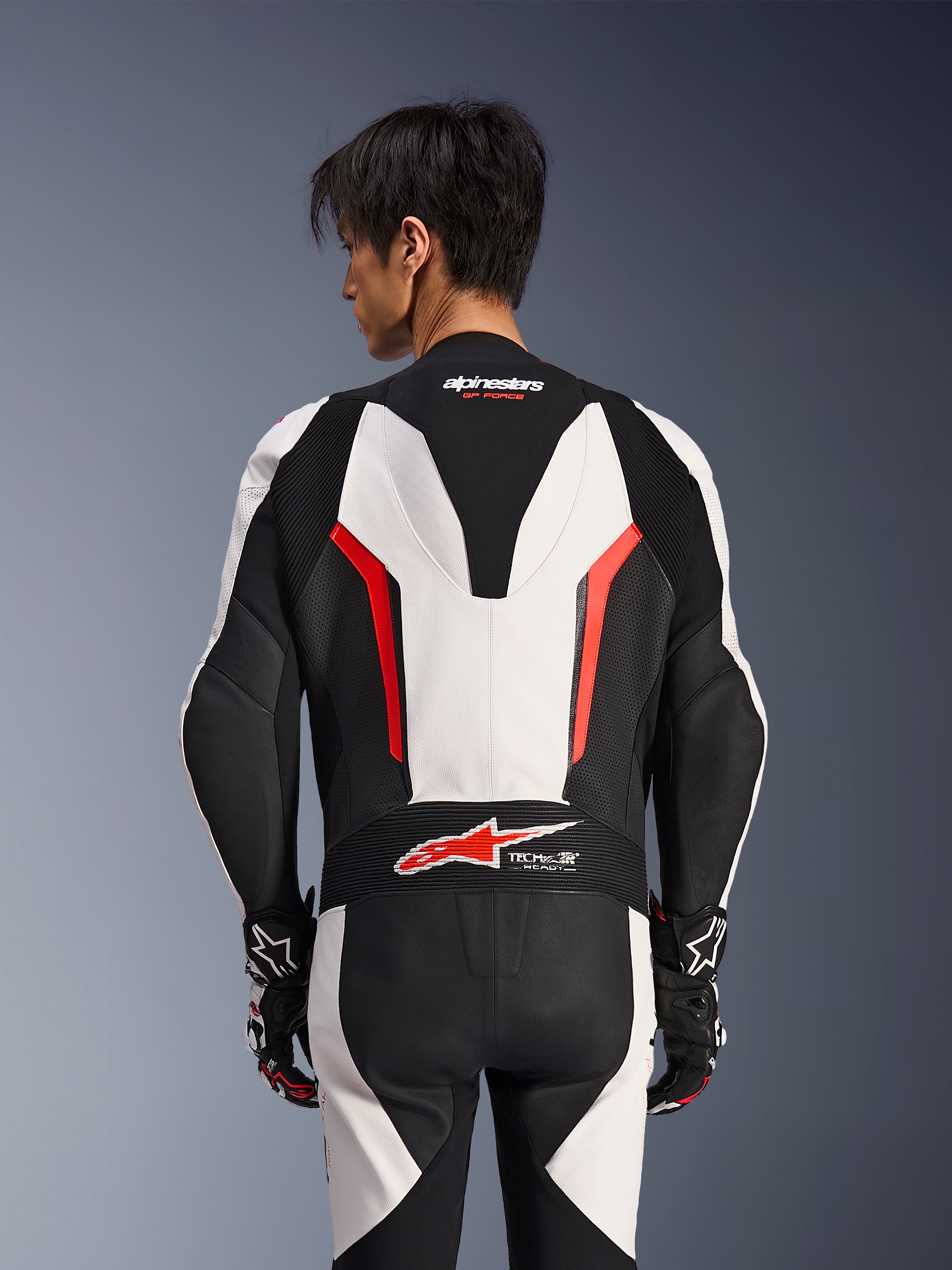 Alpinestars GP Force V2 läderställ i ett stycke, vit, svart och röd fluo, vy bakifrån på manlig modell, med aerodynamisk speedpuckel, perforerade läderpaneler och Tech-Air Ready-logotyp på nedre ryggen, professionell racingutrustning med svarta och vita handskar