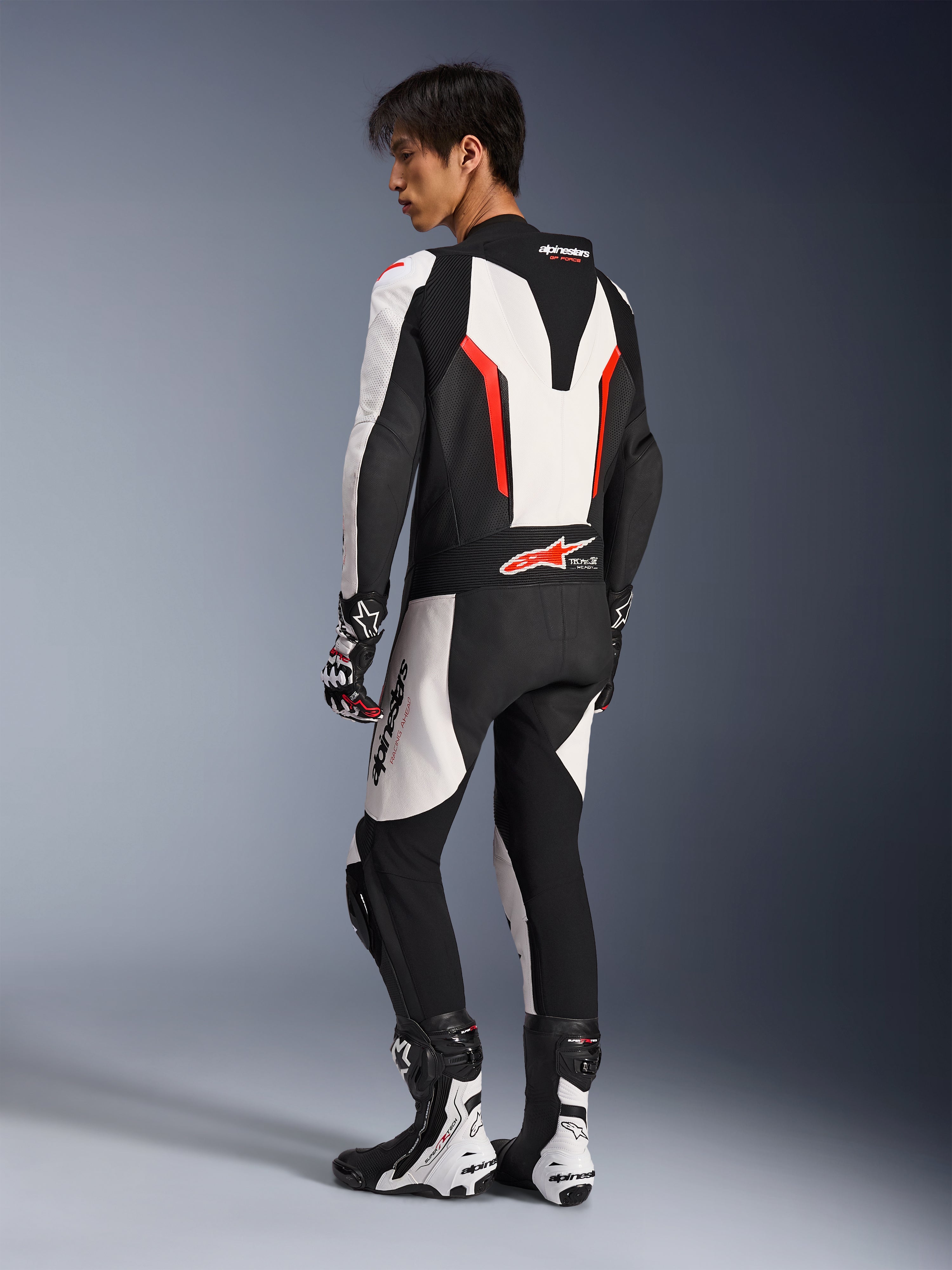 Motorcykelförare i Alpinestars GP Force V2 Leather Suit - 1Piece i vitt, svart och rött fluo, vy från sidan och bakifrån som visar den aerodynamiska puckeln och perforerade läderpaneler, matchat med racingstövlar och handskar för bana- och prestandakörning.