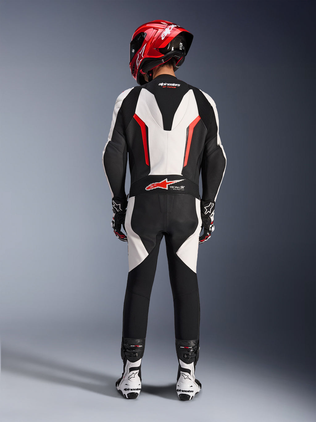 Alpinestars GP Force V2 Leather Suit - 1Piece, White Black Red Fluo, vy bakifrån som framhäver den aerodynamiska puckeln och Tech-Air Ready-märkningen, bärs med röd racinghjälm och vita och svarta stövlar, banfokuserat läderställ för prestandakörning.
