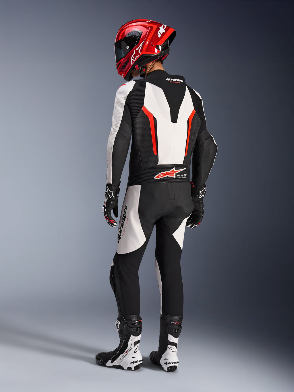 Alpinestars GP Force V2 läderdräkt - 1-dels, vit svart röd fluo, baksida av förare i full tävlingsutrustning med röd hjälm, svarta och vita stövlar och handskar, framhäver den aerodynamiska speedpuckeln och ergonomiska stretchpaneler.