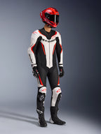 Alpinestars GP Force V2 läderställ i ett stycke i vit, svart och röd fluo, helkroppsbild i studio av en förare i komplett racingutrustning inklusive röd hjälm, svarta handskar och svarta stövlar, med aerodynamisk konstruktion i nötläder, tydlig logotyp och integrerade knäpuckar.