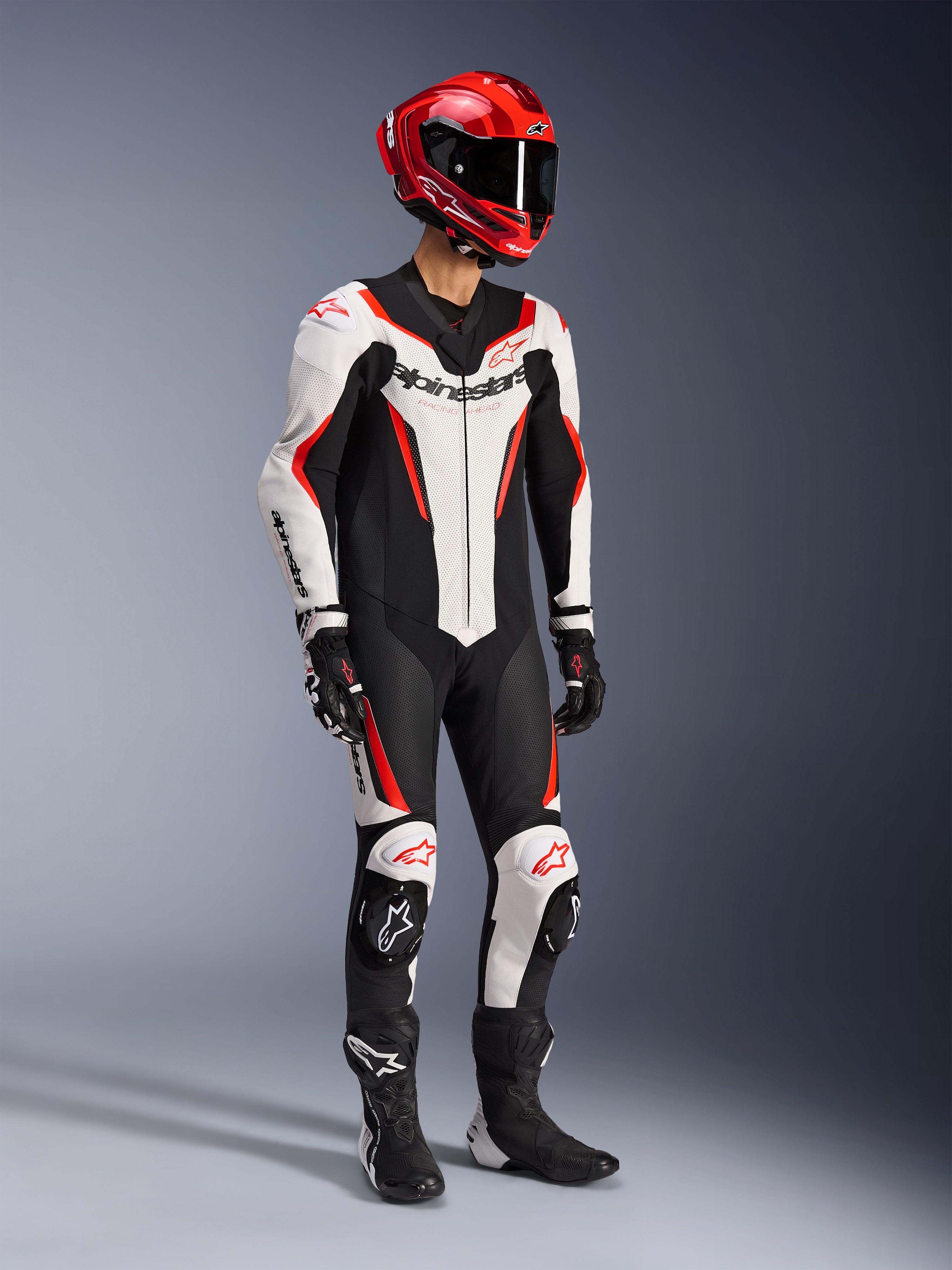 Alpinestars GP Force V2 läderställ i ett stycke i vit, svart och röd fluo, helkroppsbild i studio av en förare i komplett racingutrustning inklusive röd hjälm, svarta handskar och svarta stövlar, med aerodynamisk konstruktion i nötläder, tydlig logotyp och integrerade knäpuckar.