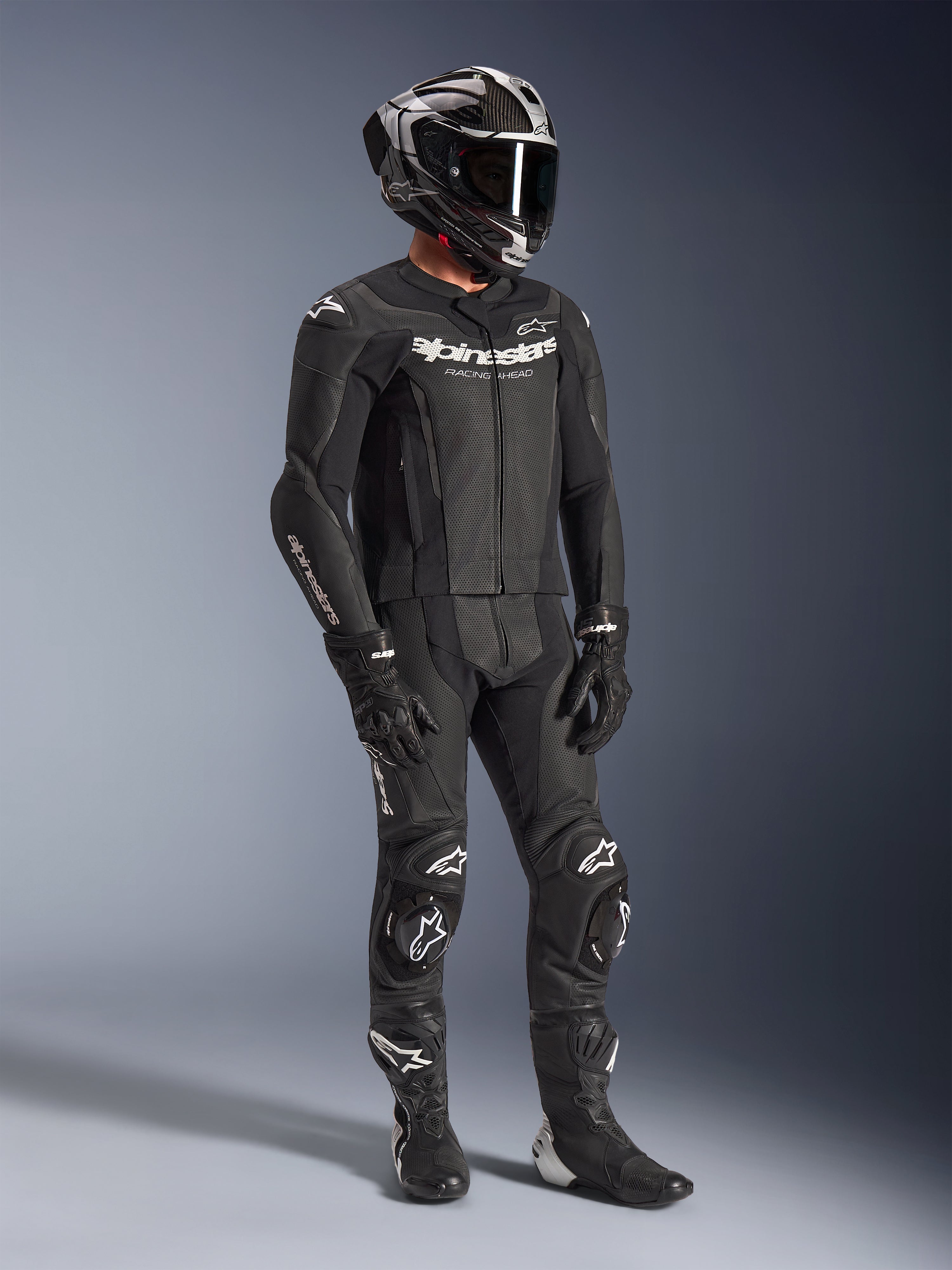 Alpinestars GP Force V2 2-Piece Leather Suit, black motorcycle racing suit, worn with a full-face helmet, gloves, and boots, featuring perforated leather construction, aerodynamic speed hump, and white Alpinestars logos on the chest, arms, and knees.