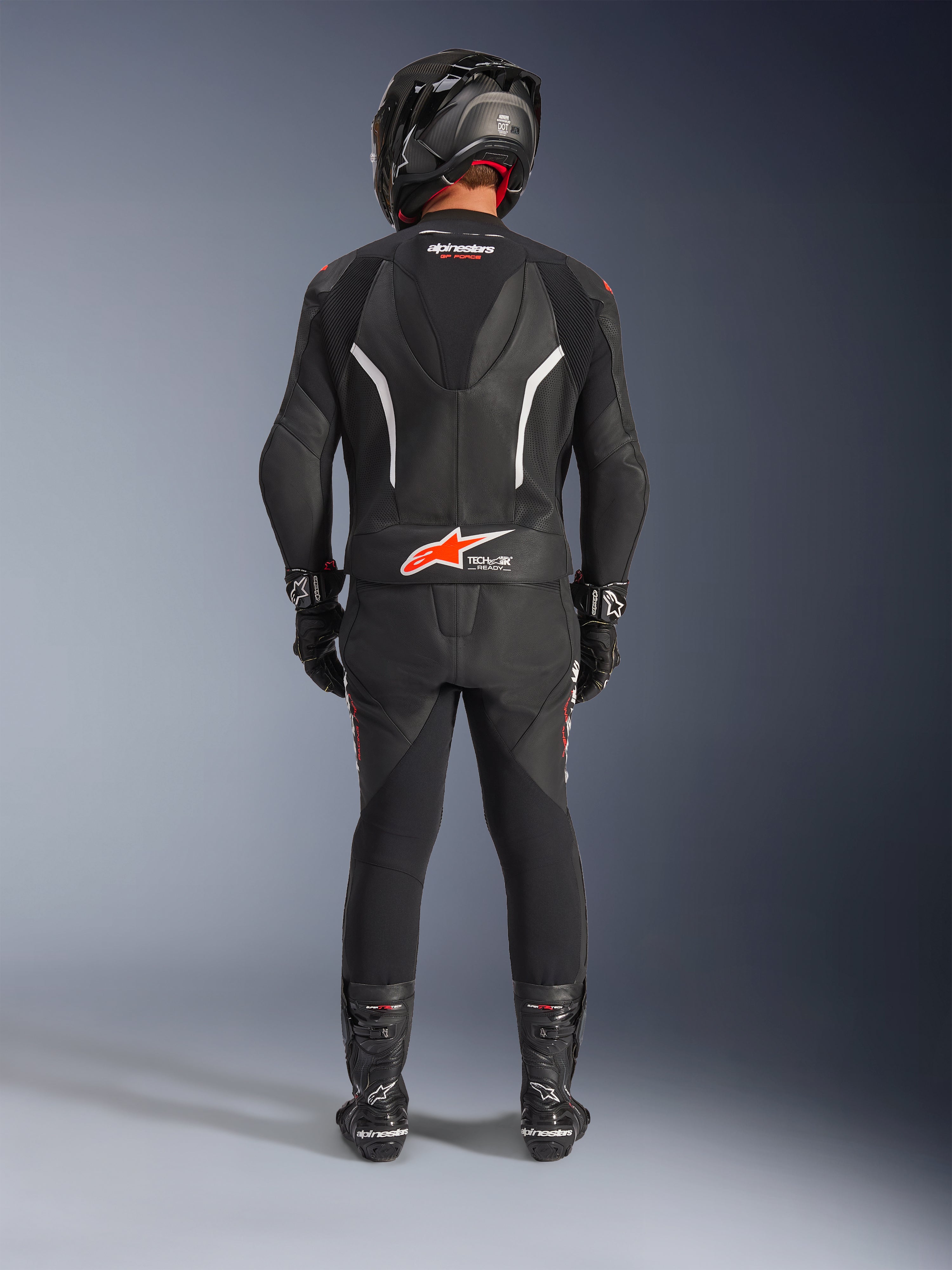 Motorcycle rider from behind wearing the Alpinestars GP Force V2 2-Piece Leather Suit, black with white and red fluo accents, featuring an aerodynamic hump and Tech-Air Ready logo, complete with a black racing helmet, gloves, and boots.