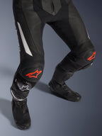 Alpinestars GP Force V2 2-Piece Leather Suit, motorcycle racing pants in black with red and white accents, featuring perforated leather, accordion stretch panels above the knees with red star logos, worn with black motorcycle boots, high-performance racing gear