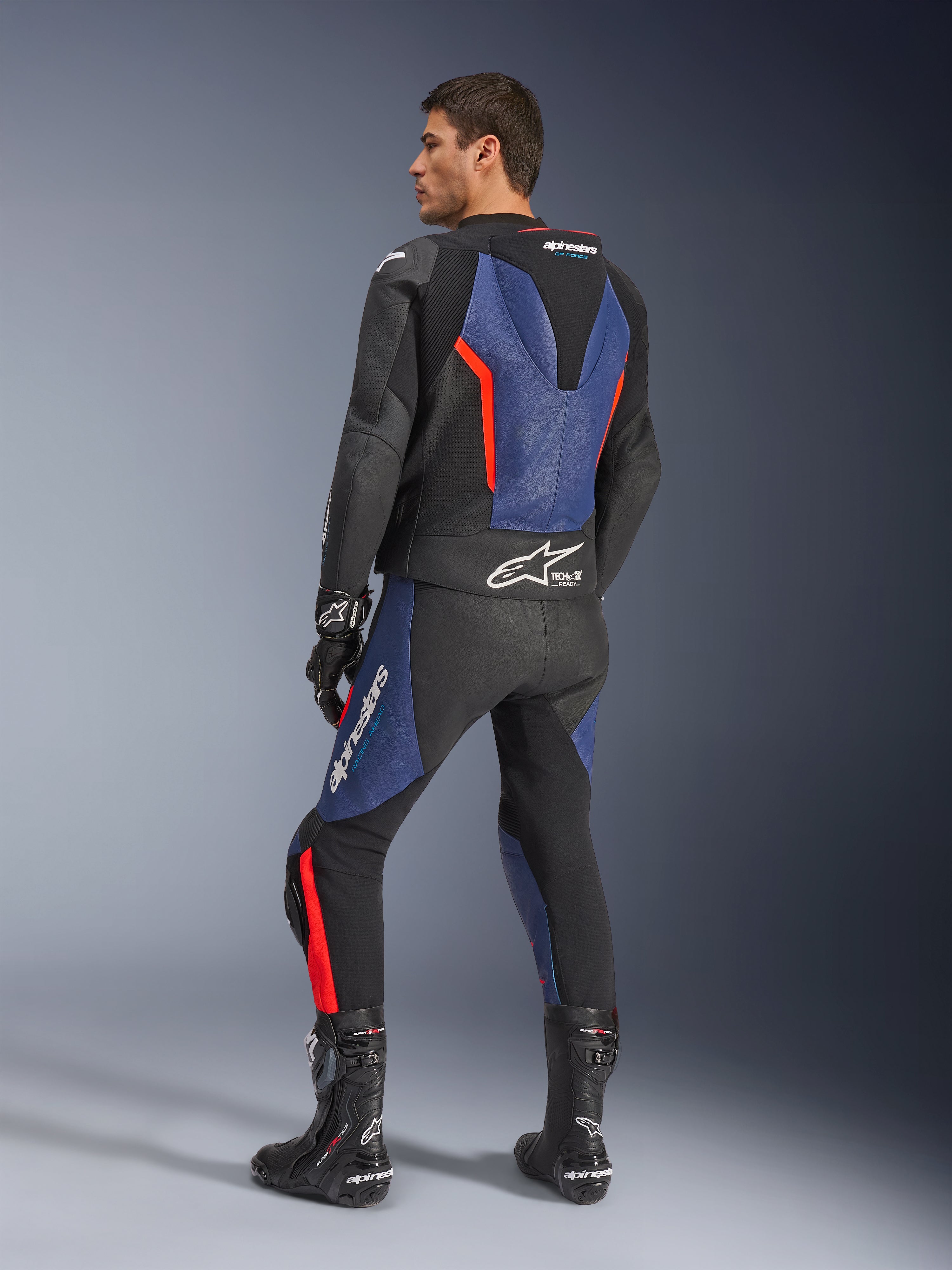 Alpinestars GP Force V2 läderställ i två delar, svart, blå och fluorescerande röd, profilvy bakifrån på en modell, med aerodynamisk puckel, perforerat läder och Tech-Air Ready-märkning, bärs med svarta Alpinestars racingstövlar och handskar
