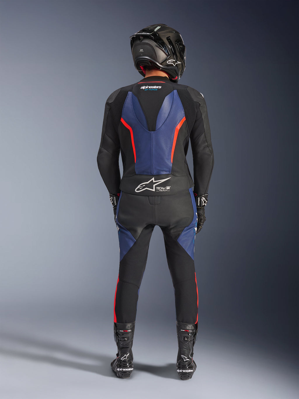 Alpinestars GP Force V2 Läderställ 2-delat, Black Blue Red Fluo, fullständig vy bakifrån av en förare i komplett racingutrustning, med blå läderpaneler på ryggen och benen med fluorescerande röda accenter, Tech-Air® Ready-logotyp på nedre ryggen, tillsammans med en kolfiberhjälm, svarta handskar och racingstövlar.