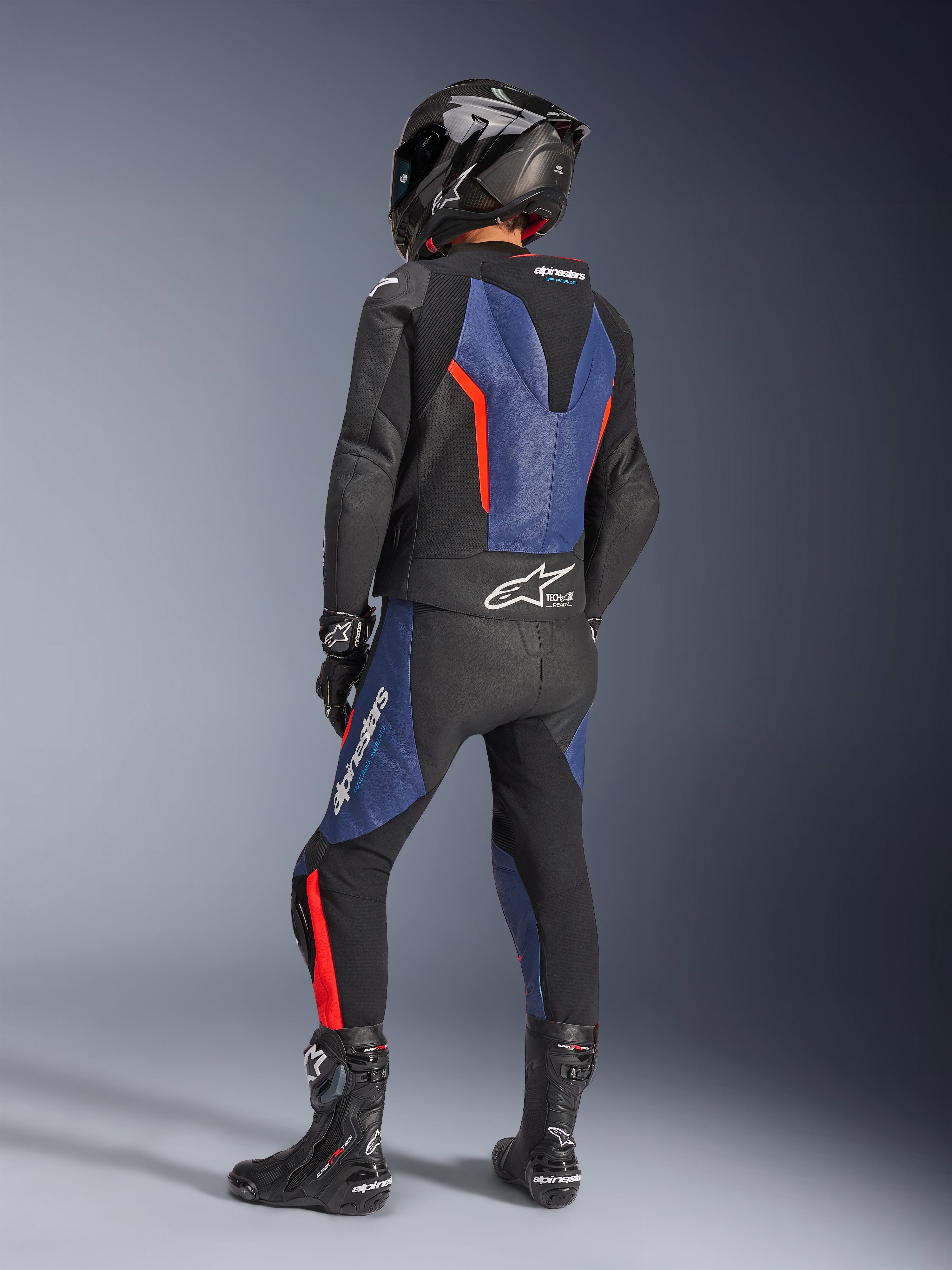 Alpinestars GP Force V2 tvådelat läderställ, Black Blue Red Fluo, vy bakifrån av förare i professionell racingutrustning med integrerad speedpuckel och Tech-Air Ready-märkning, blå och röd fluo-detaljer på svart läder, bärs med kolfiberhjälm och svarta stövlar, högpresterande motorcykelutrustning