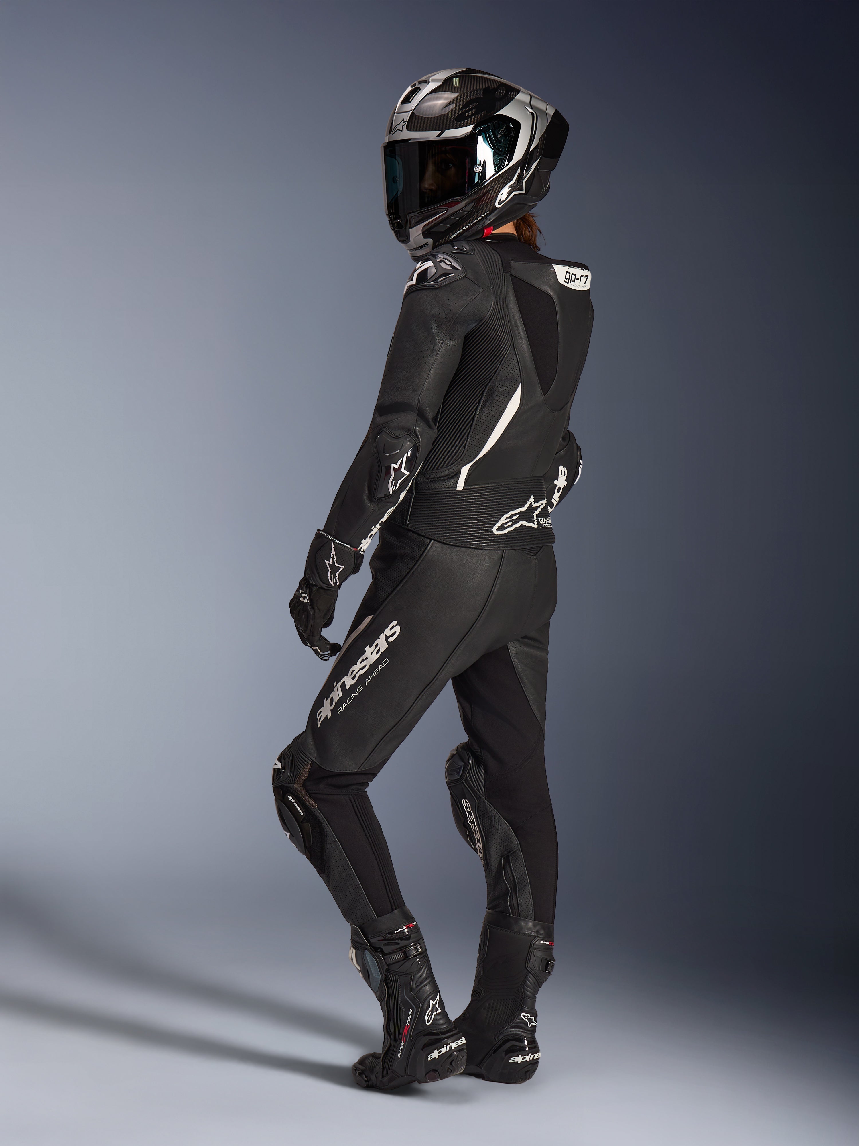 Kvinnlig förare i Alpinestars Stella GP-R7 2-delat läderställ i svart, integralhjälm för racing med kolfiberfinish, matchande handskar och stövlar, ryggvy som visar den aerodynamiska puckeln och vita Alpinestars-logotyper, professionell utrustning för bana och sportkörning