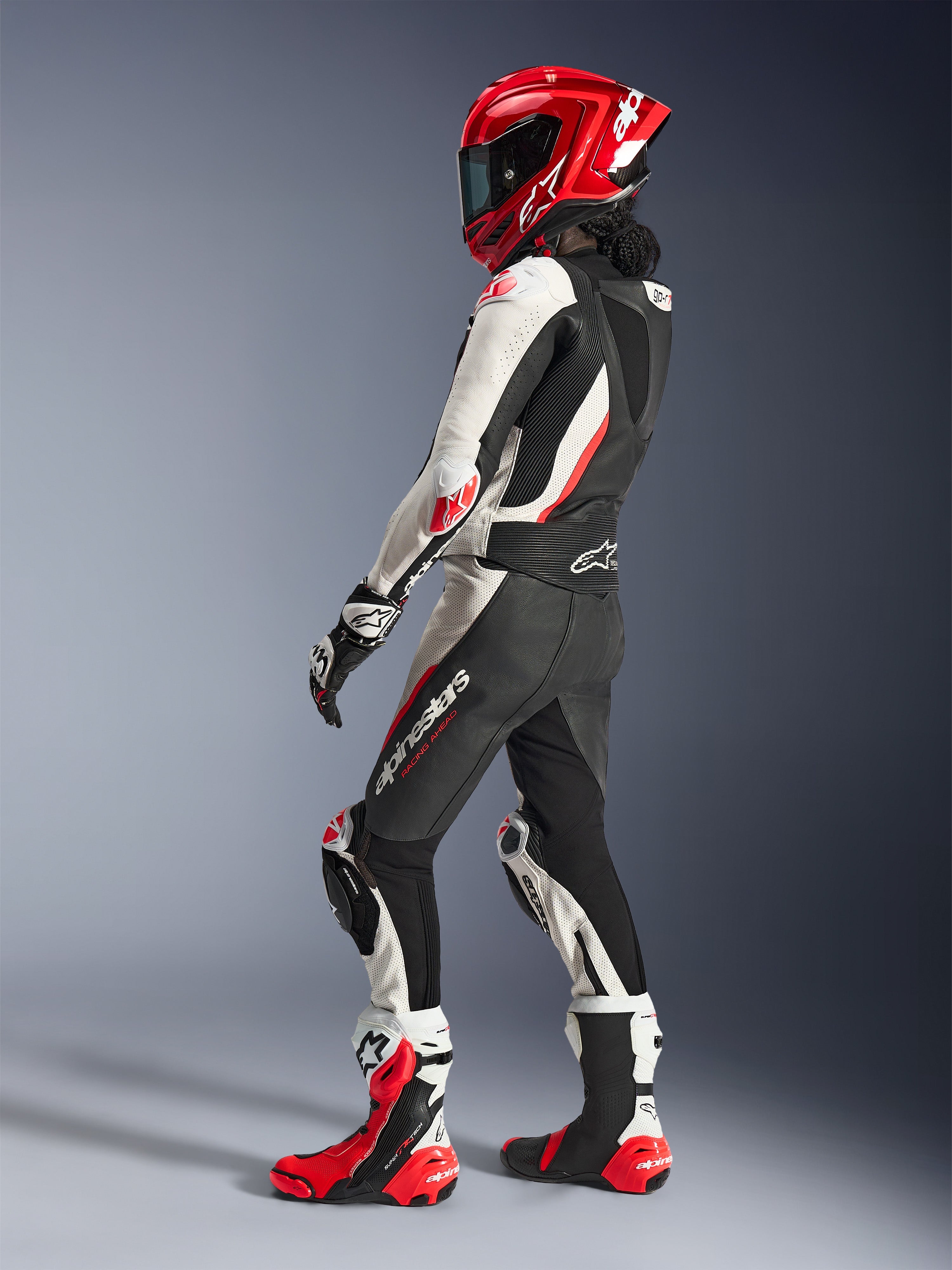 Kvinnlig förare klädd i Alpinestars Stella GP-R7 2-delat läderställ i svart, vitt och röd fluo, sedd bakifrån i trekvartsvy som visar den aerodynamiska puckeln, matchande röd integralhjälm, svarta och vita racinghandskar samt röda och vita tekniska racingstövlar, professionell racingutrustning.