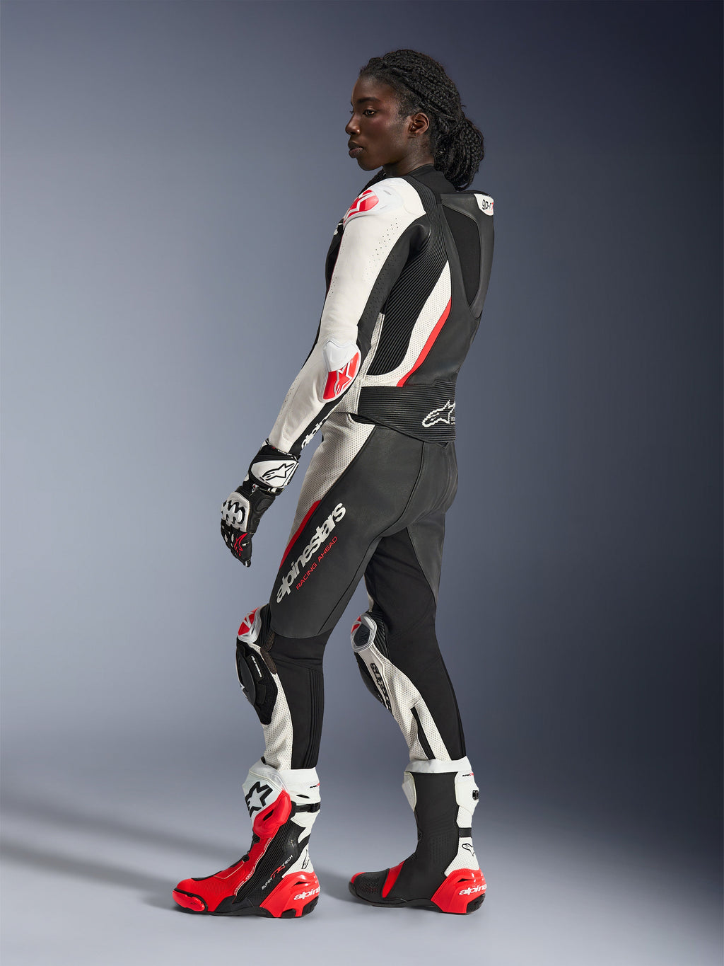 Kvinna i Alpinestars Stella GP-R7 2-delat läderställ, svart, vit och röd fluo, sidoprofil som visar aerodynamisk puckel och knäsliders, matchande Alpinestars racinghandskar och stövlar, professionell motorcykelutrustning för bana
